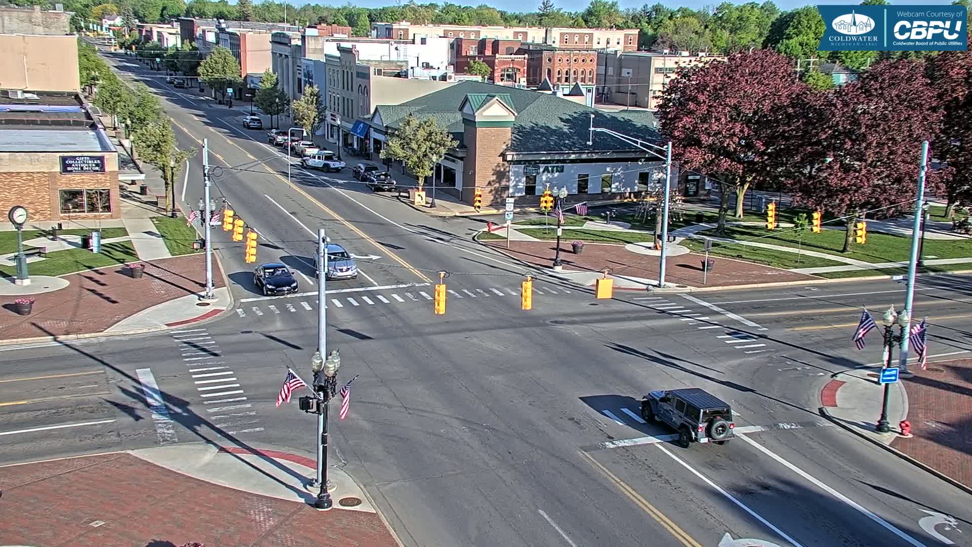 A sunny day reveals a town square intersection with several cars, a few pedestrians, and lush green trees surrounding a brick-paved area.