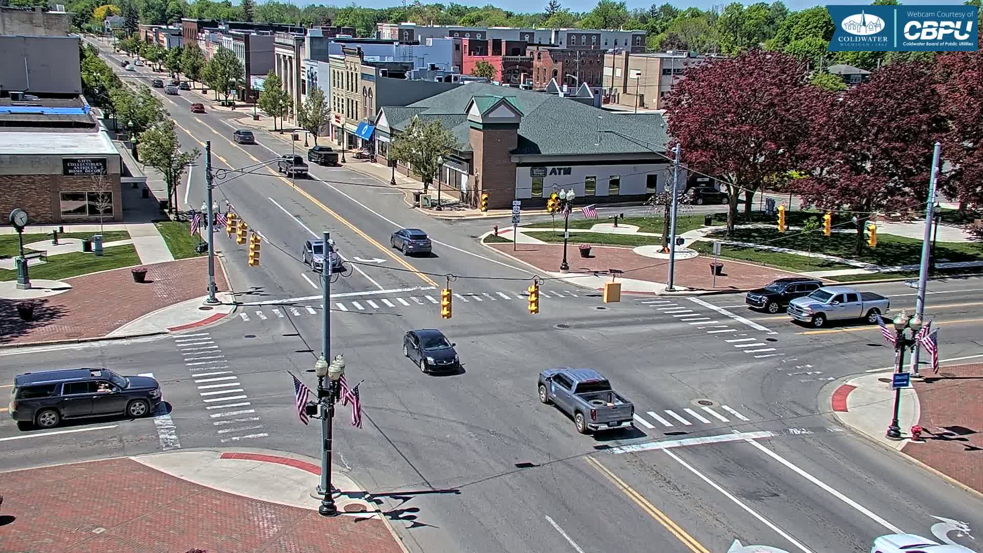 A sunny day shows a town's intersection with several cars, brick sidewalks, and leafy green trees.