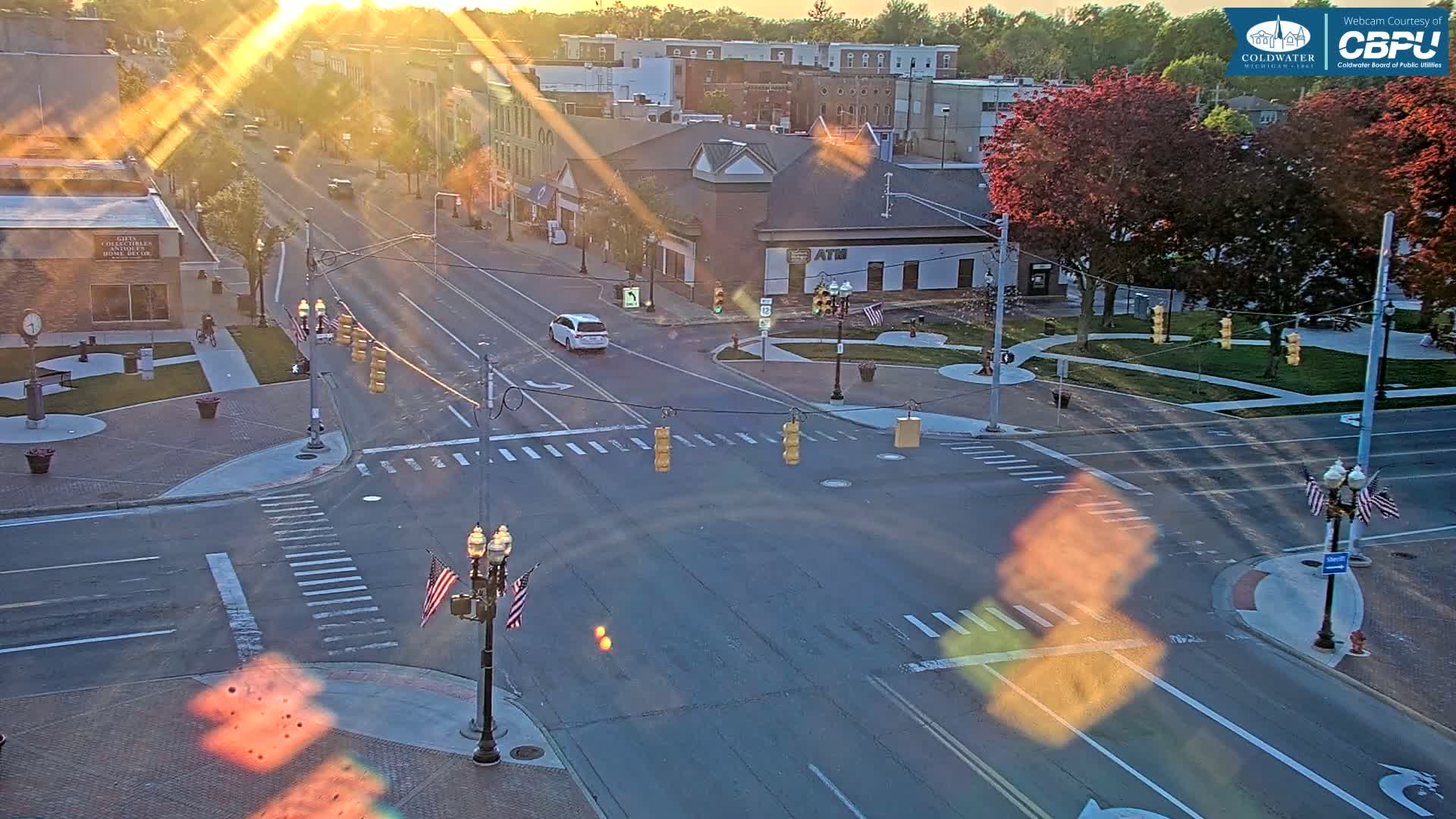 A sunlit, high-angle view of a town intersection with traffic lights, a small park, and several buildings, showing a mostly clear sky.