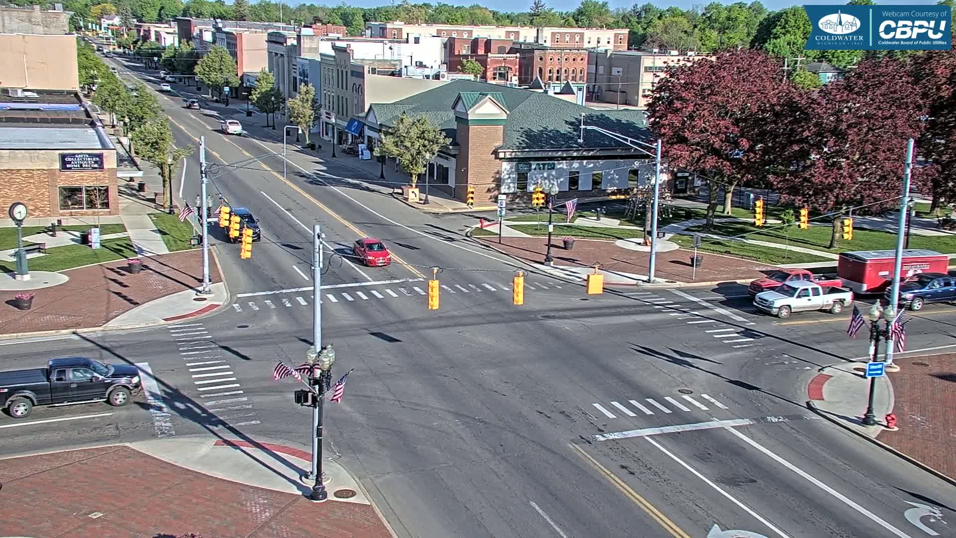 A sunny day reveals a mostly empty street intersection in a town, with several vehicles at the crosswalk and buildings lining the street.