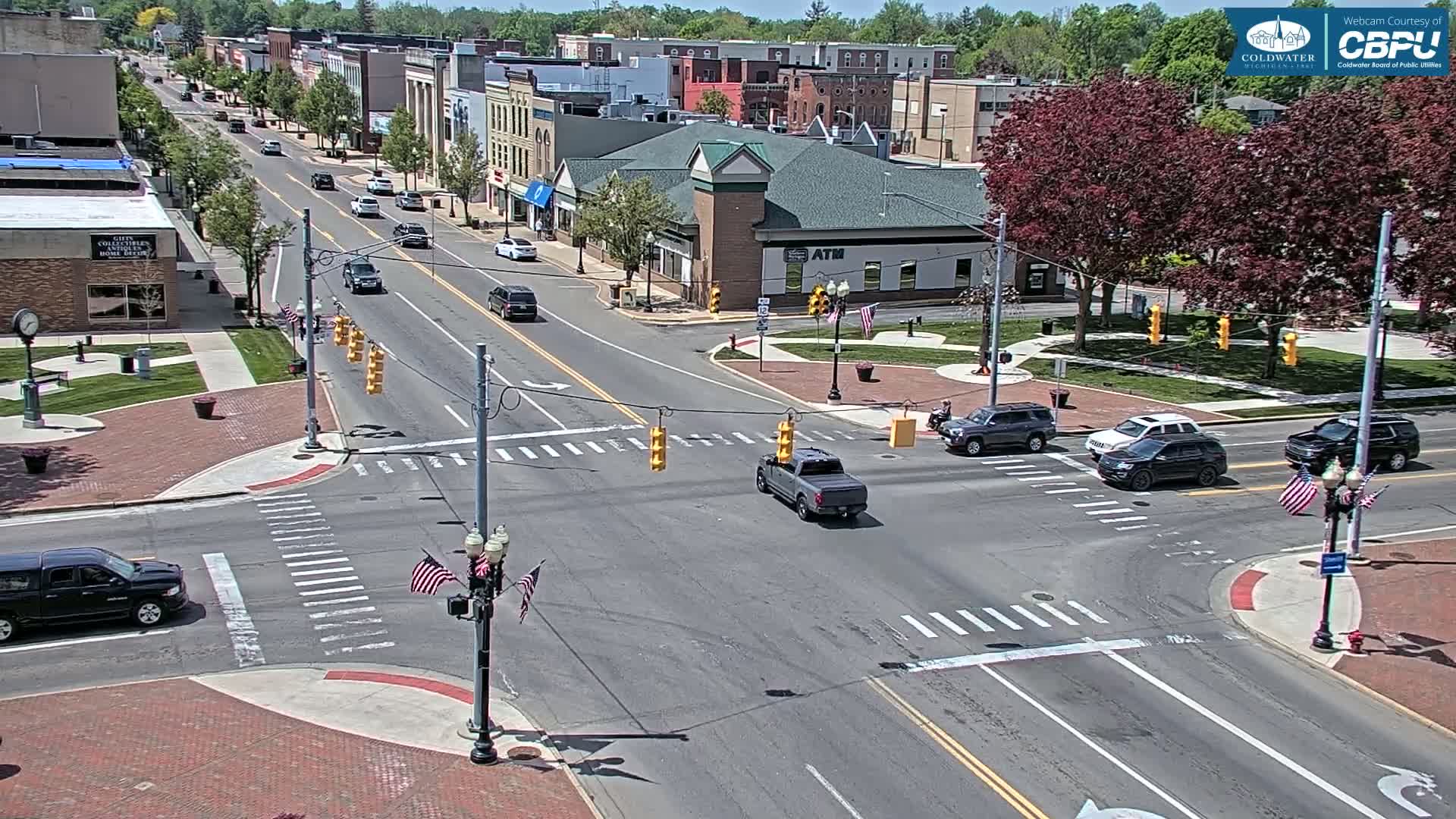 A sunny day shows a city intersection with several cars, brick sidewalks, and green spaces.