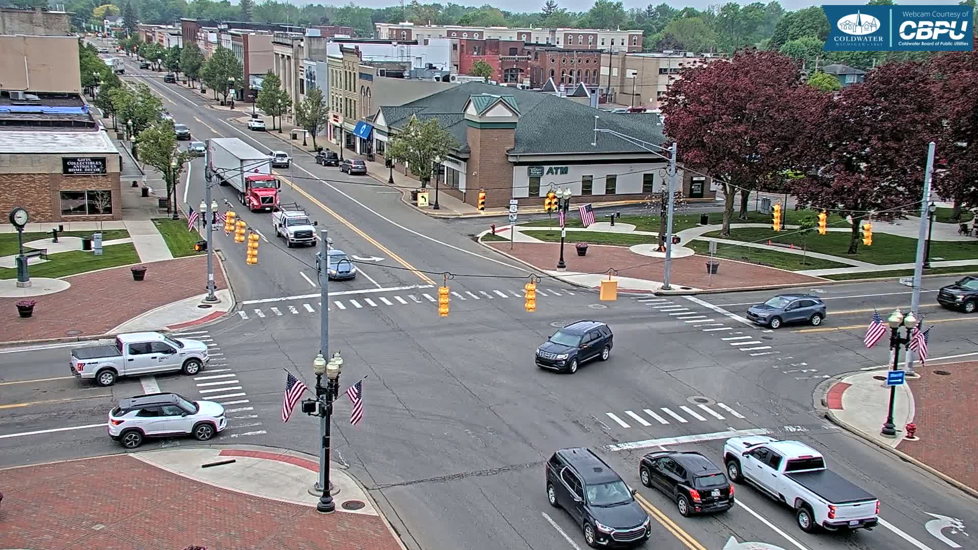 A mostly sunny day shows a high-angle view of a town's intersection with several vehicles at a four-way stop, surrounded by brick sidewalks, green spaces, and buildings.