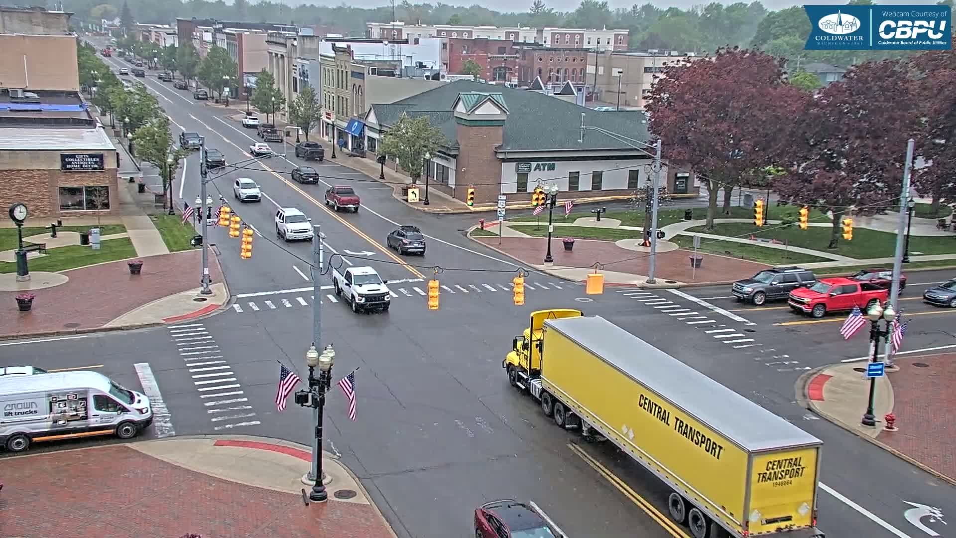 A yellow semi-truck travels through a town intersection on a wet, overcast day, with several other vehicles and pedestrians visible.