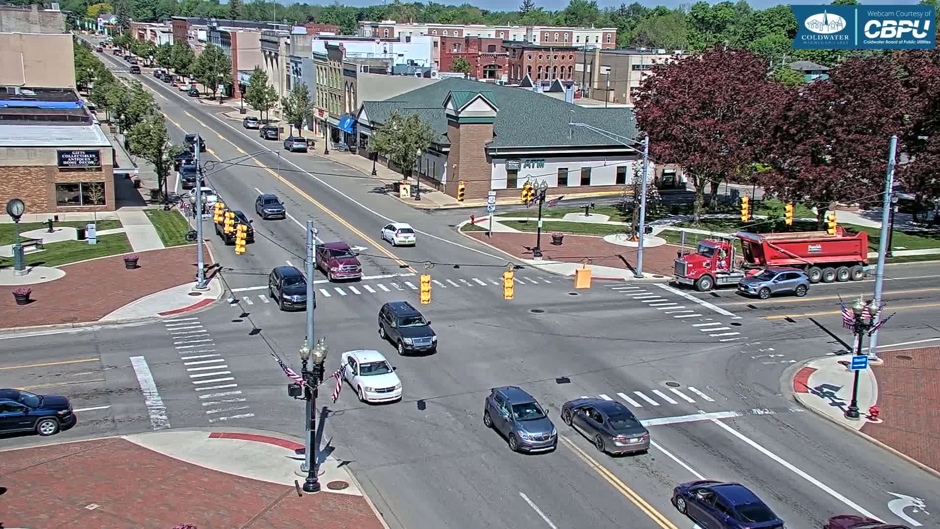 A sunny day in a town square with several vehicles at a traffic intersection, surrounded by brick sidewalks and buildings.