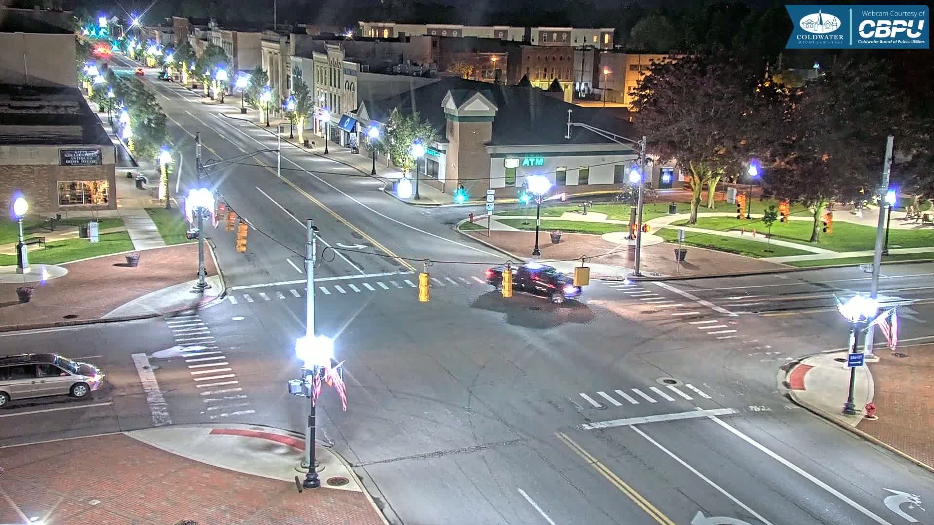 A nighttime, high-angle view of a town square intersection with several vehicles, streetlights, and buildings.