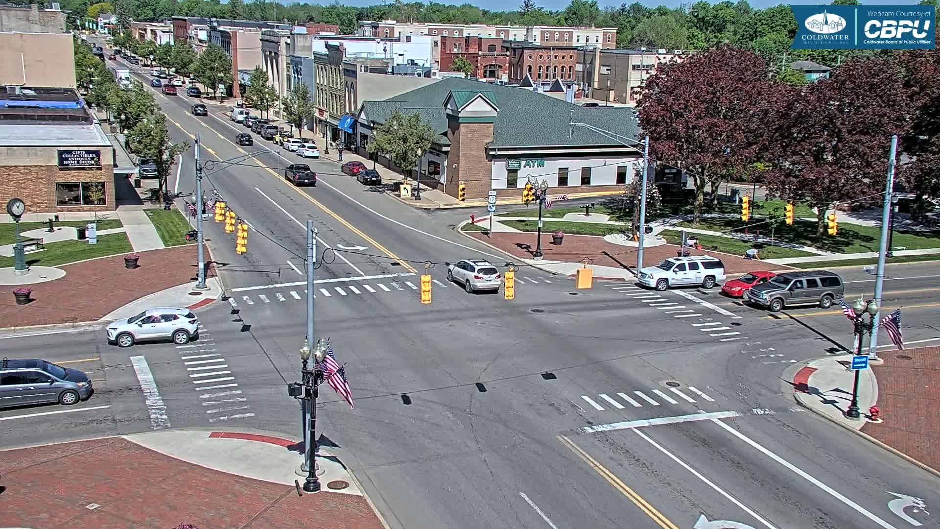 A sunny day reveals a town square intersection with several cars, brick sidewalks, and buildings lining a main street.