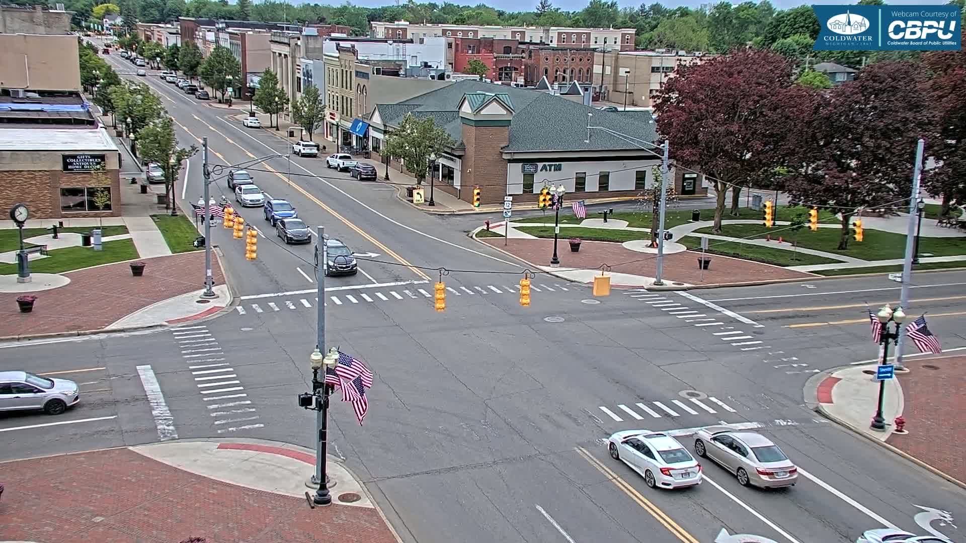 A mostly sunny day shows a small-town intersection with several cars at a four-way stop, surrounded by brick sidewalks, buildings, and green spaces.