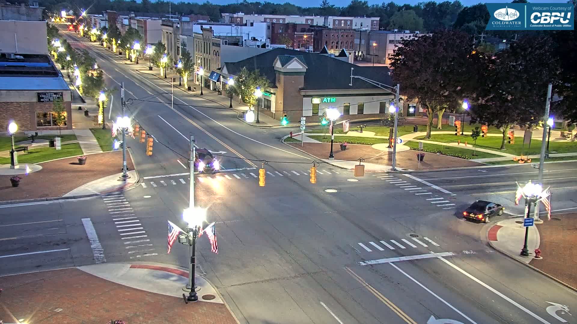 A brightly lit street intersection in a town at night, with a few cars, and trees lining the sidewalks.