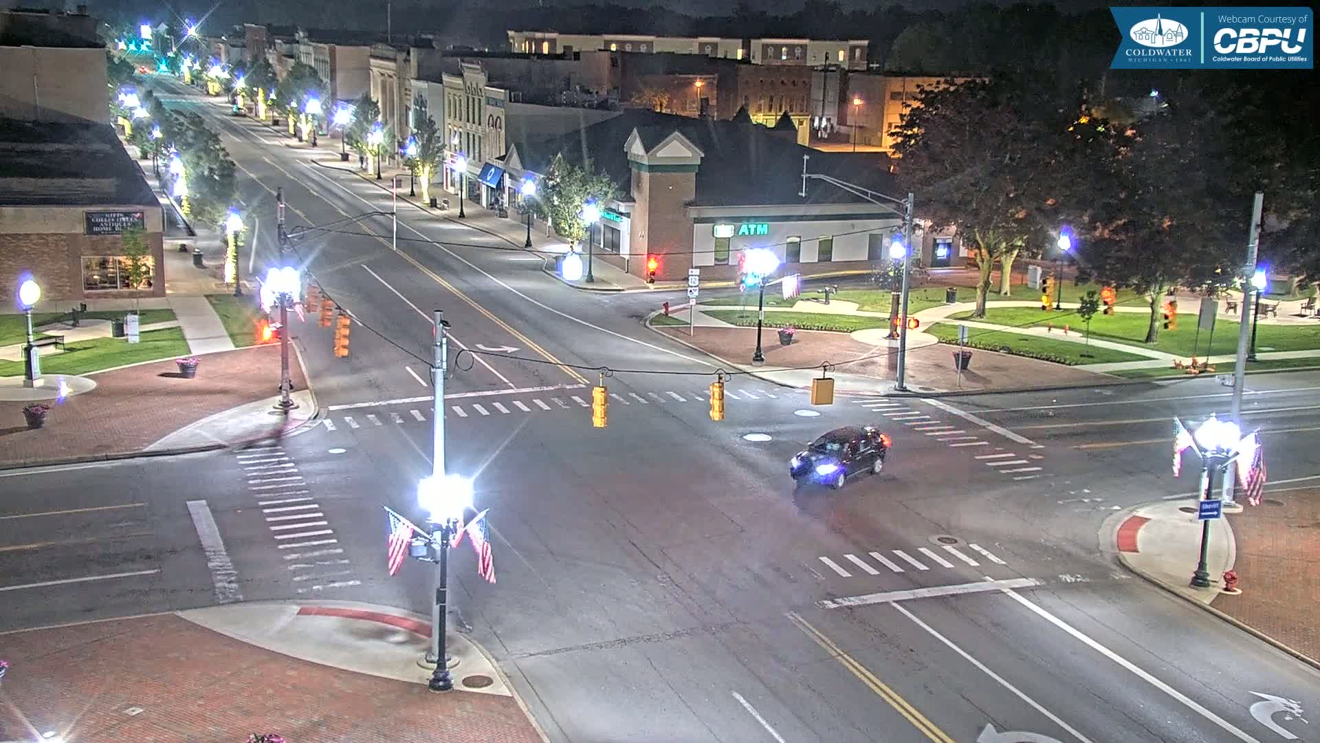 A nighttime, overhead view of a town square intersection with traffic lights, street lamps, and a few cars, under clear skies.