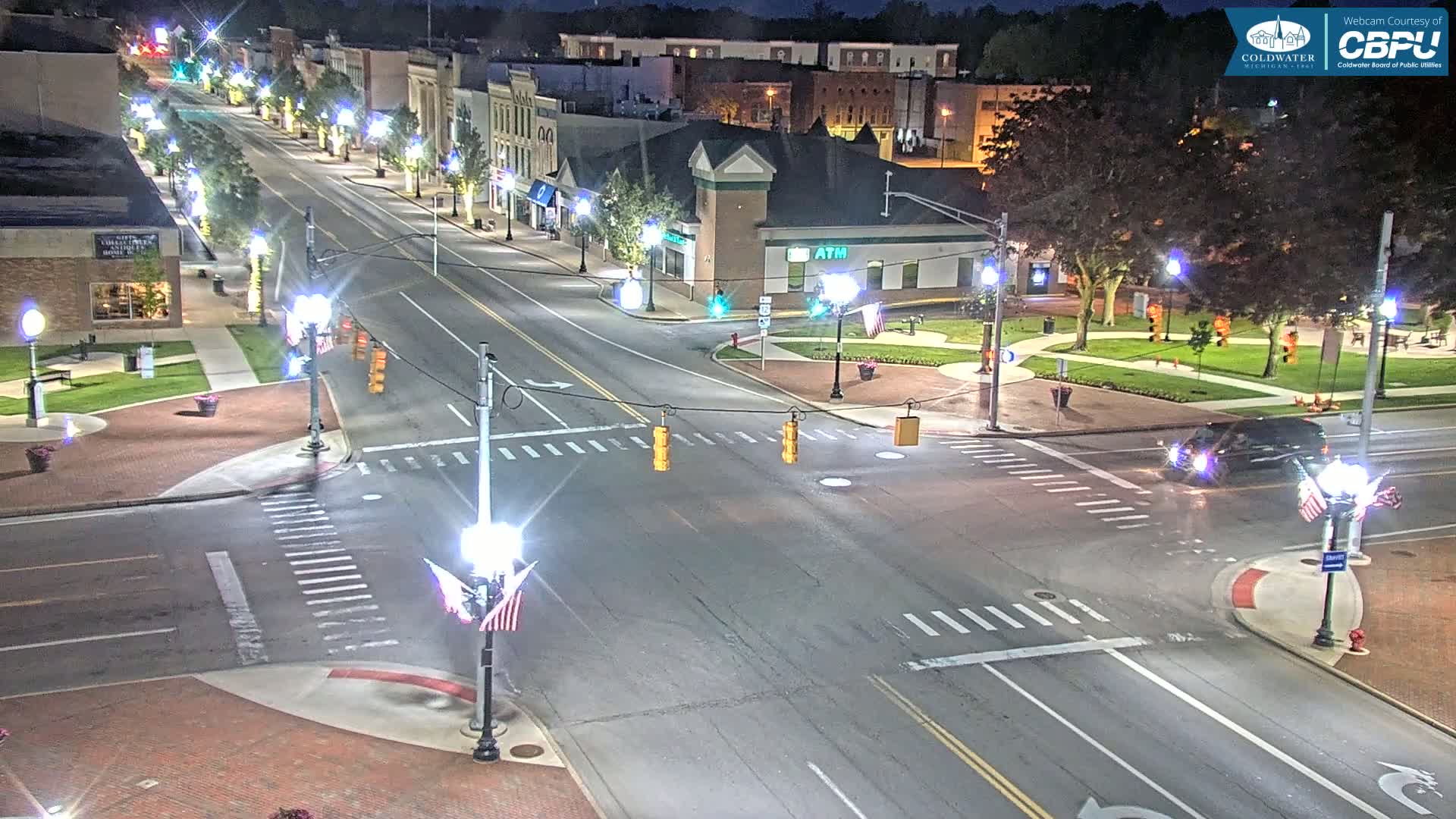 A well-lit, tree-lined street in a town at night, with a car driving through a crossroad intersection.