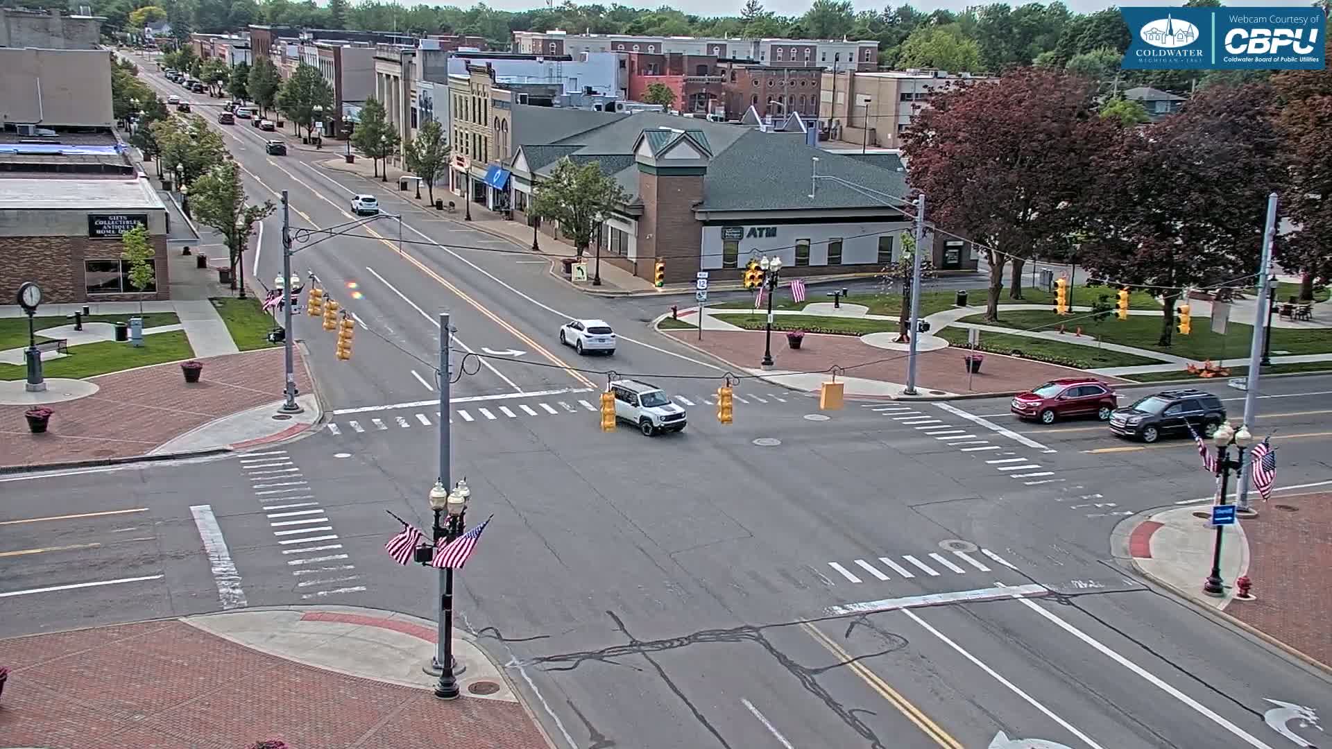 A mostly sunny day shows a small-town intersection with several cars, American flags on light poles, and brick sidewalks and streets, all surrounded by buildings and lush green trees.