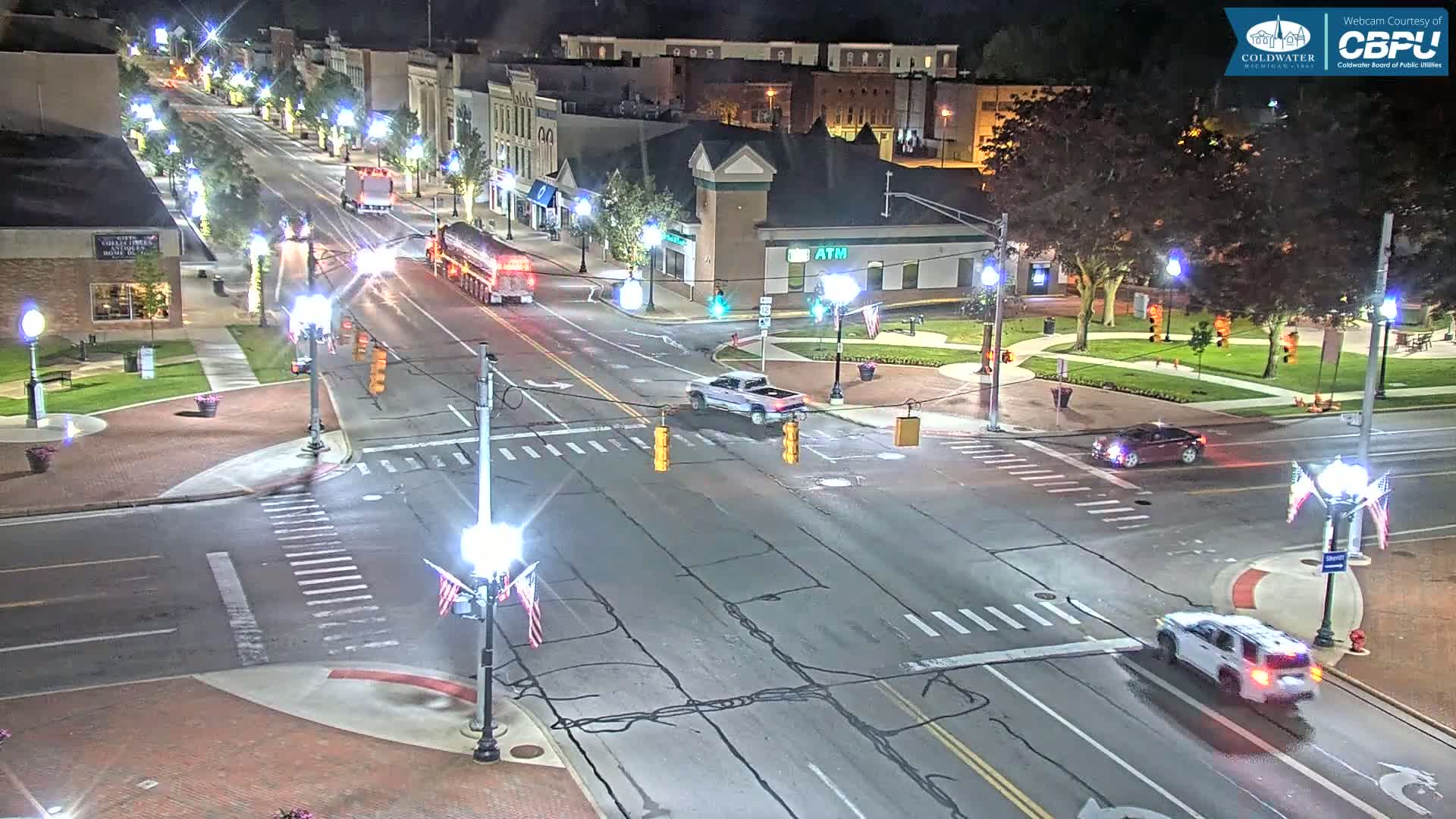 A nighttime aerial view of a town intersection with several vehicles, streetlights, and buildings.