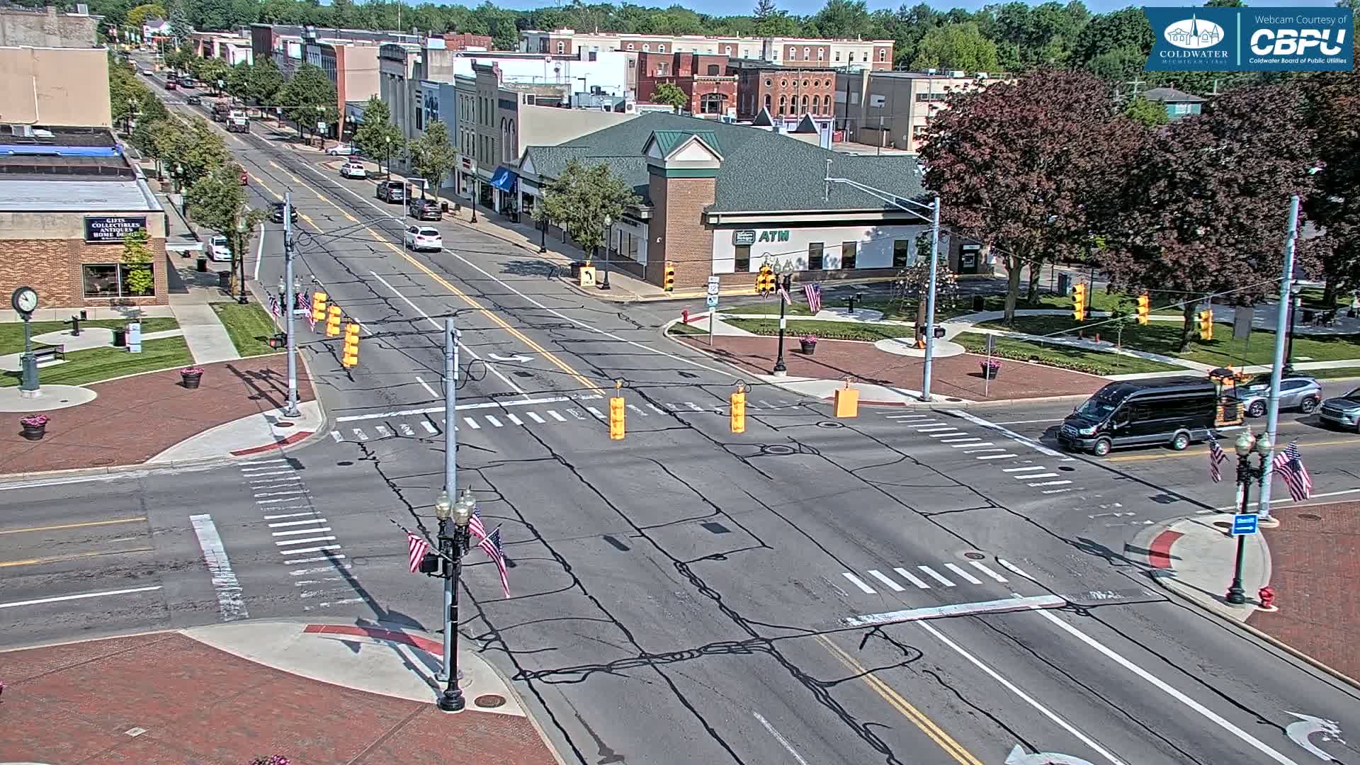 A sunny day shows a view of a town's main street intersection, with several cars and a van present, and brick sidewalks and landscaping surrounding the roads.