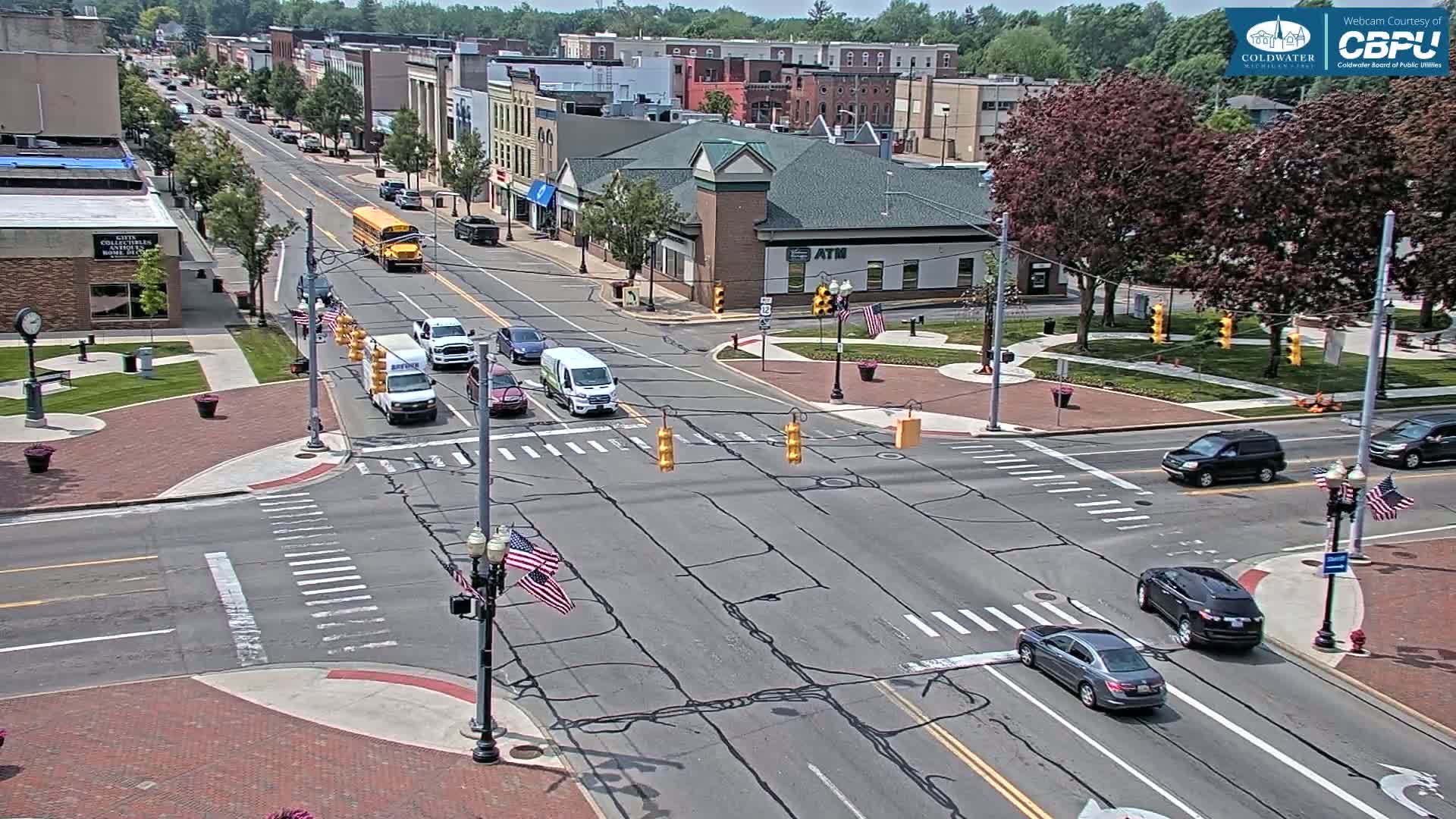 A sunny day reveals a town square intersection with several cars and a school bus, surrounded by brick buildings and lush green trees.