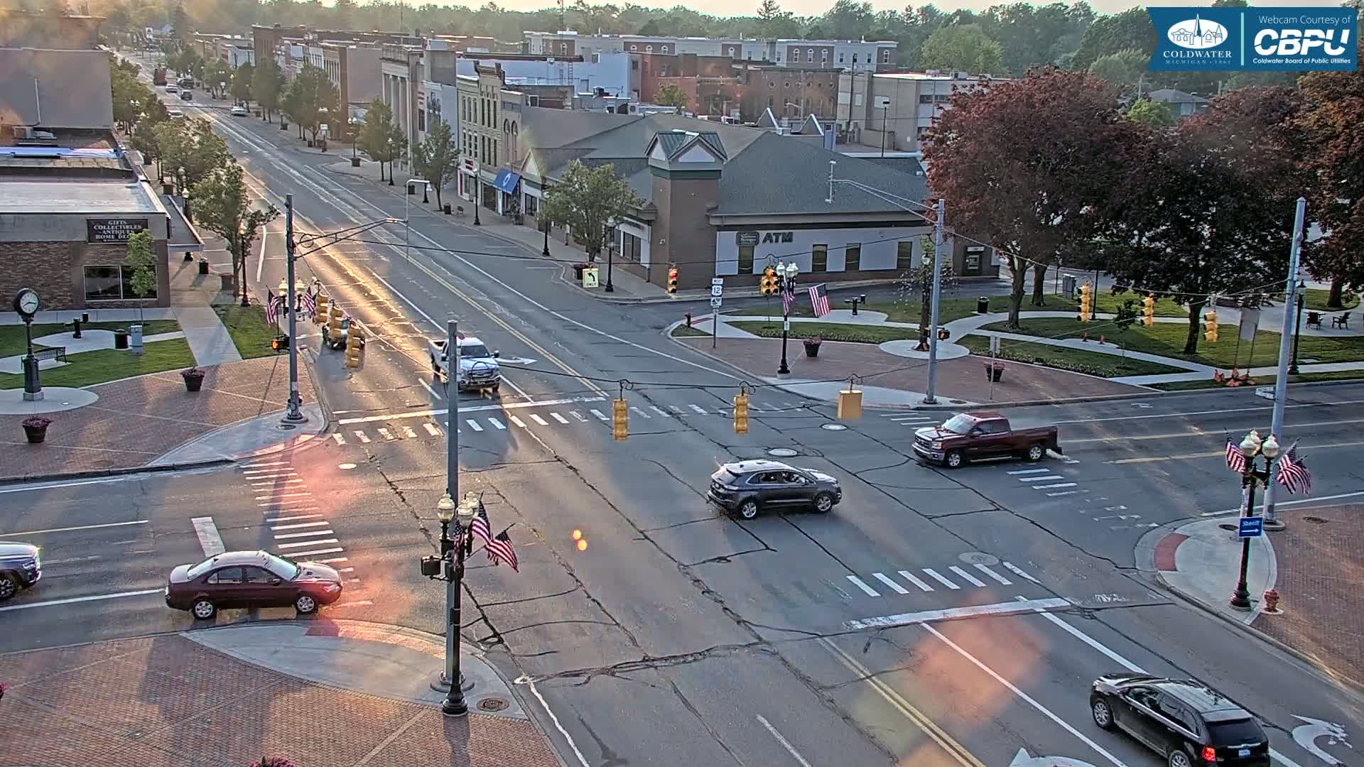 A high-angle, long shot of a city intersection on a sunny day with several cars driving through,  surrounded by buildings and trees.