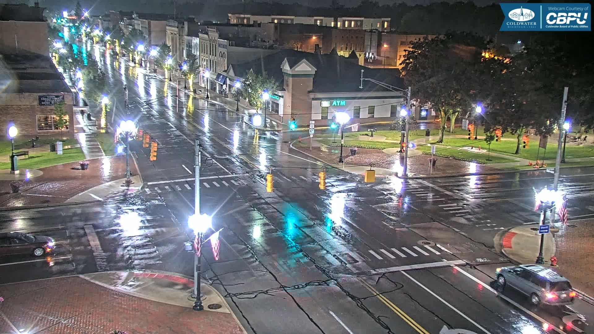 A rain-slicked town square at night, illuminated by streetlights, shows several cars driving through a wet intersection.