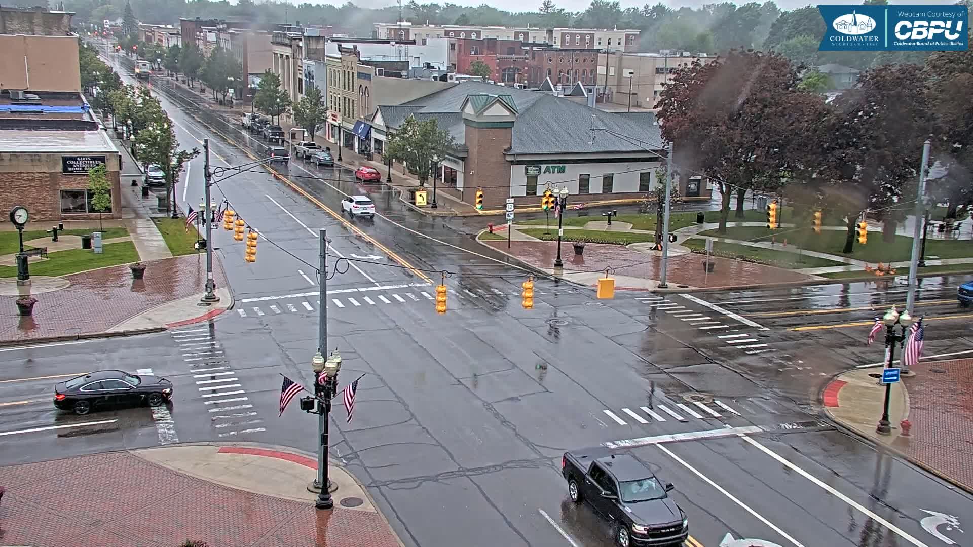 A rain-slicked city intersection shows several cars at a traffic light, with buildings and trees visible in the background.