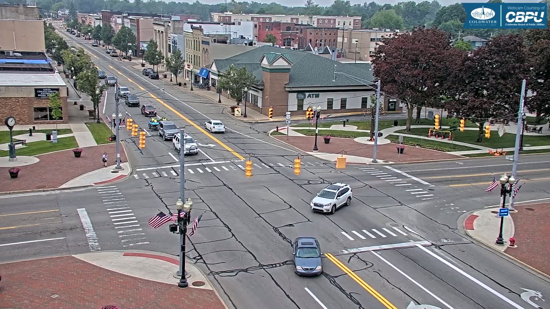 A mostly sunny day shows a town intersection with several cars driving, a pedestrian walking, and American flags waving near a light pole.