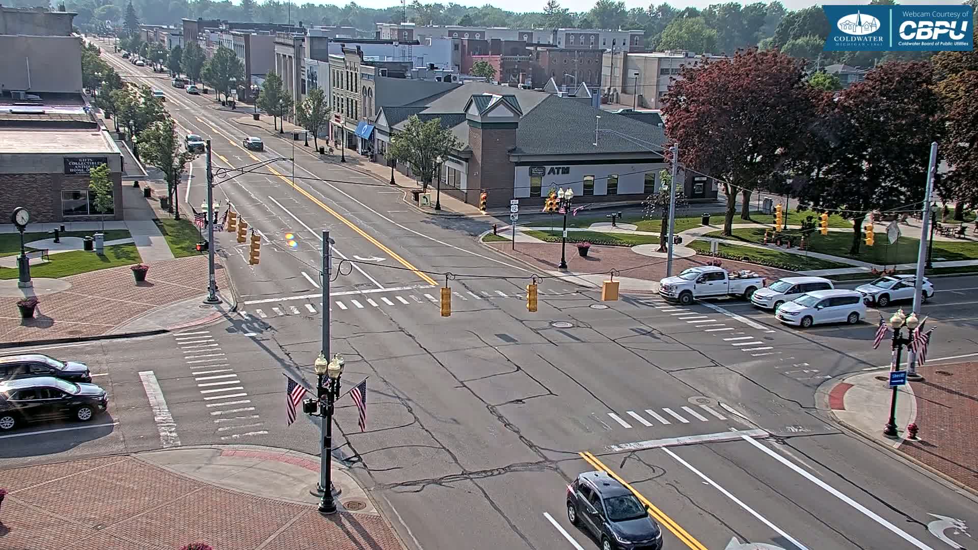 A sunny day reveals a city street intersection with several cars, surrounded by brick sidewalks, low-lying greenery, and multi-story buildings.