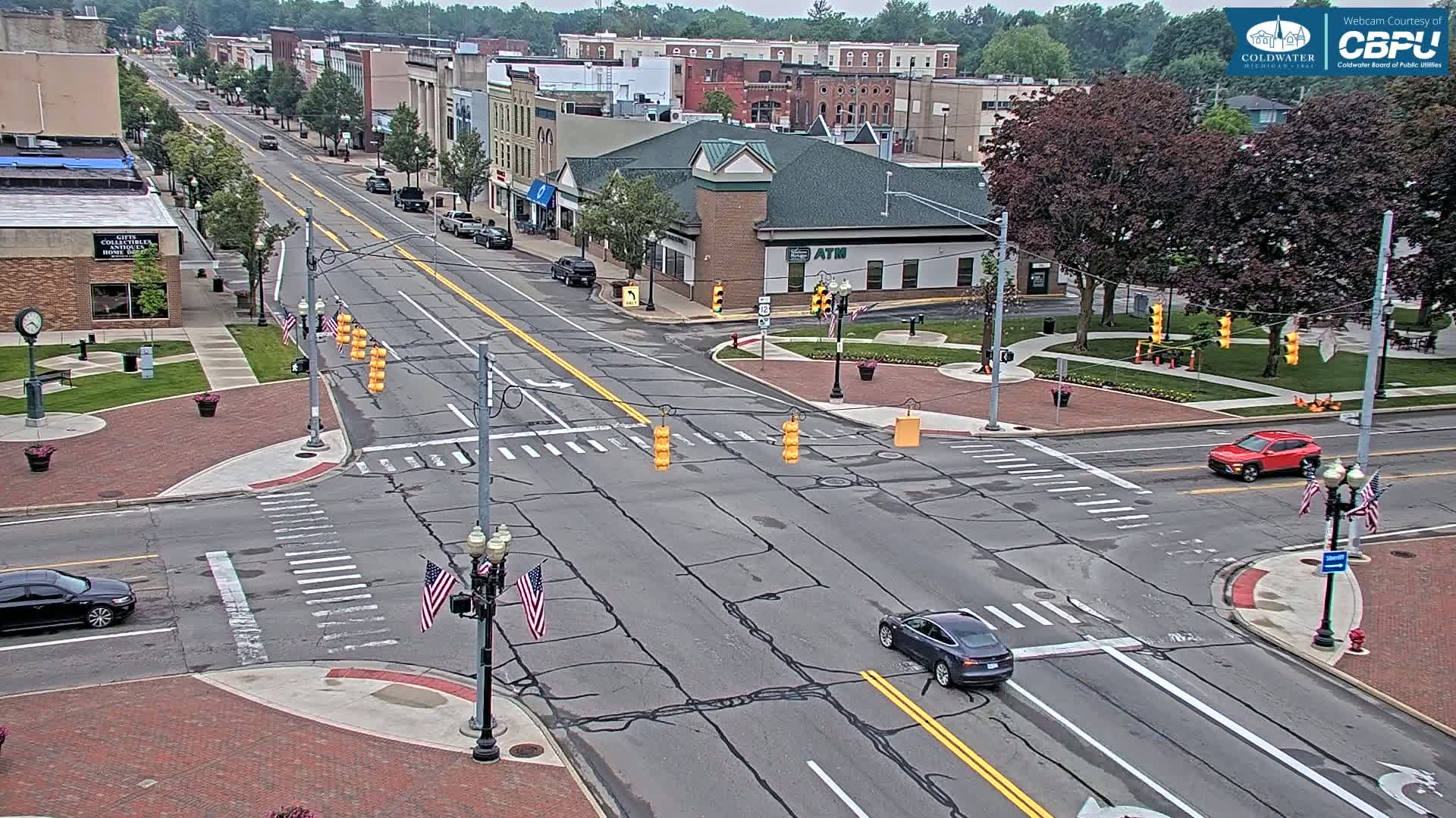 A mostly sunny day shows a street intersection in a town with several cars, buildings, and small American flags decorating light posts.