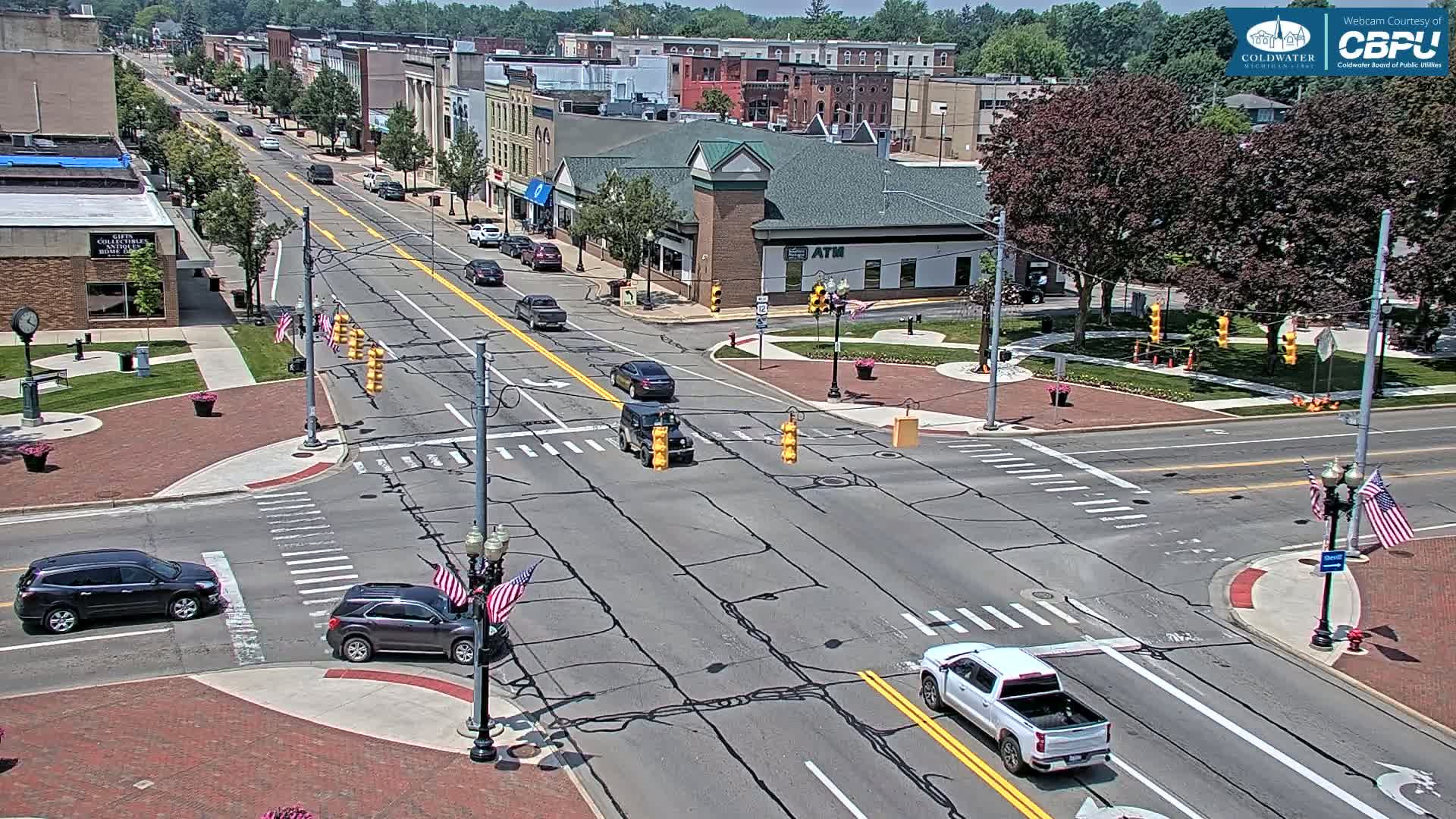 A sunny day shows a four-way intersection in a town with several vehicles, brick sidewalks, and American flags adorning light posts.