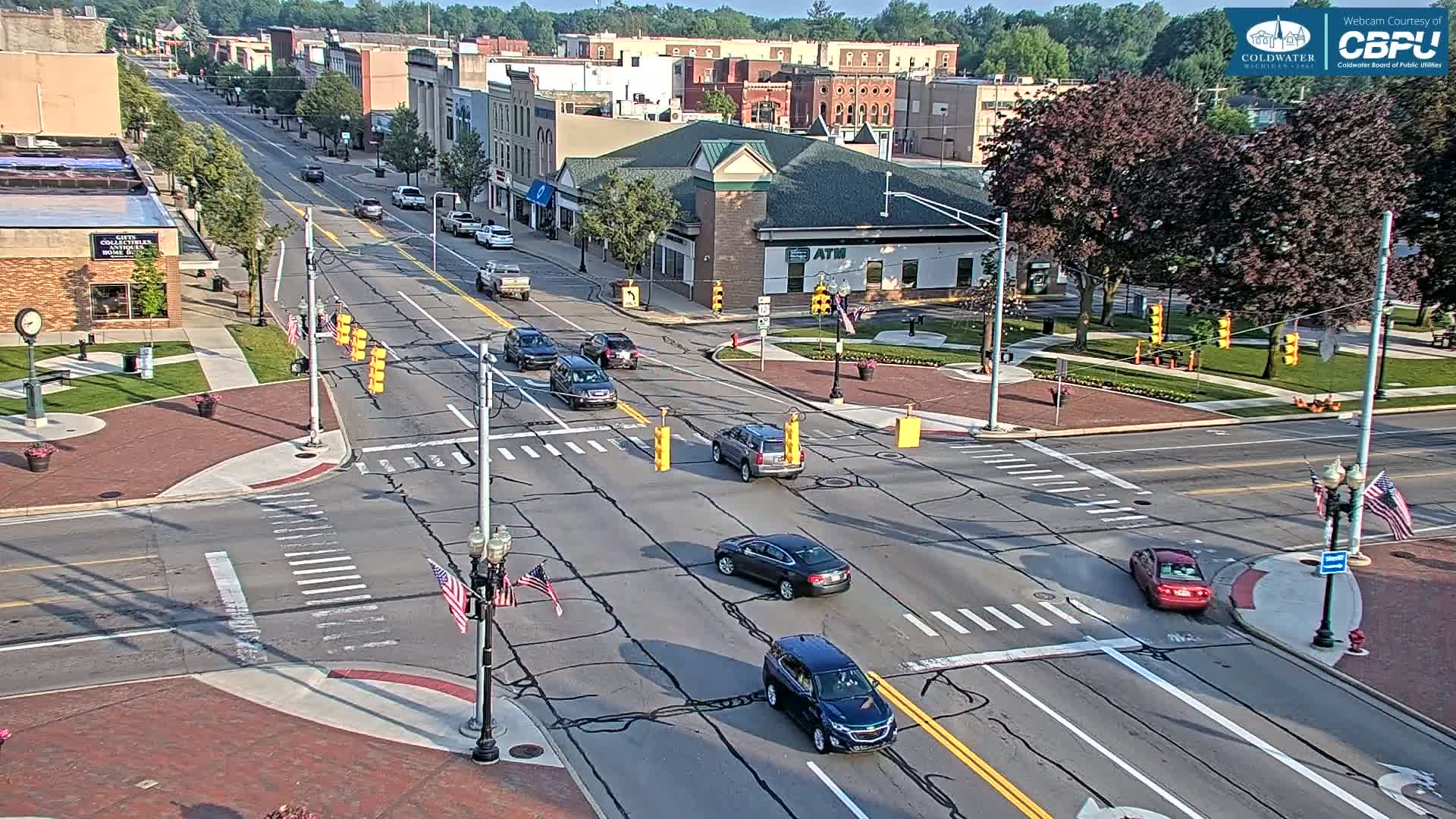A sunny day reveals a street intersection in a town with several cars driving through and buildings lining the street.