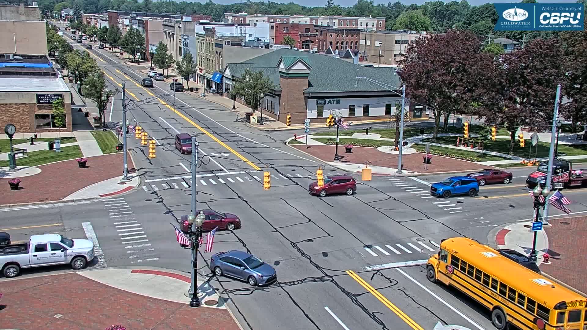 A sunny day in a town square with several cars and a school bus at an intersection in front of brick buildings.