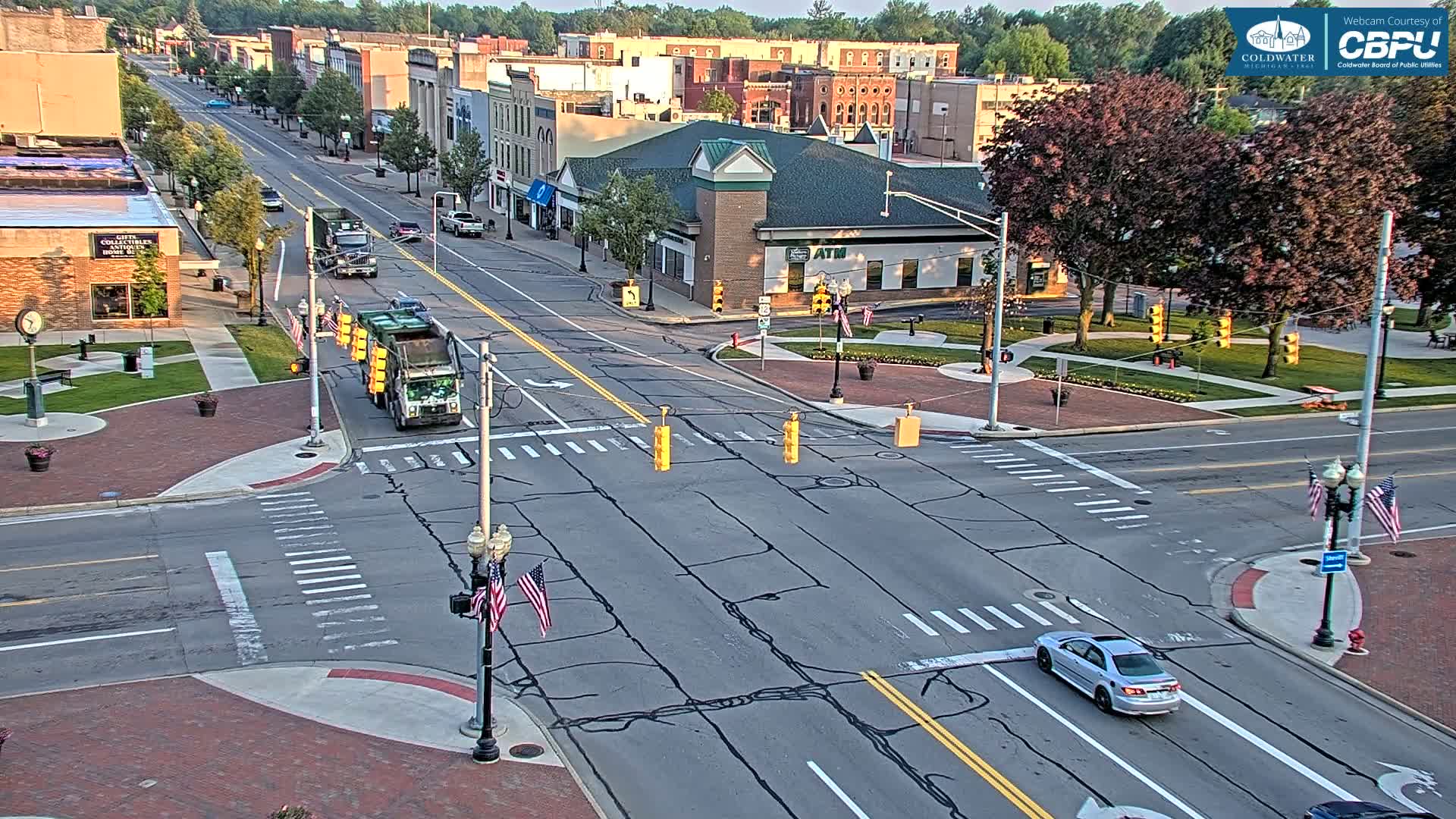 A sunny day reveals a town square intersection with two garbage trucks and a silver car; several buildings line the street.