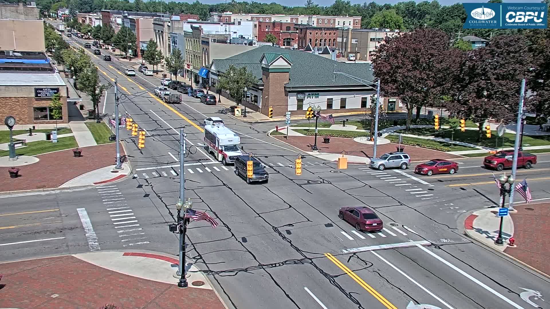 A sunny day reveals a town square intersection with several vehicles and pedestrians, surrounded by brick buildings and lush green trees.