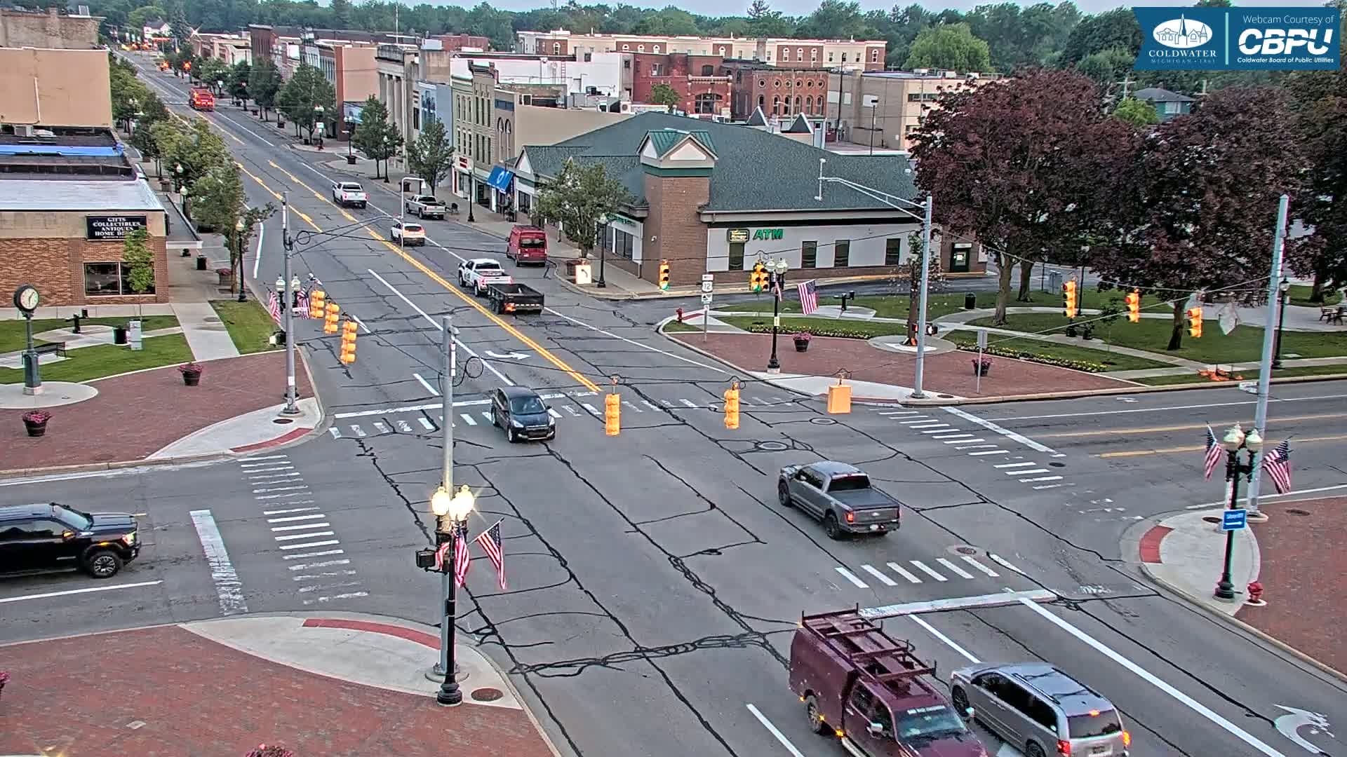 A mostly empty street intersection in a town, under partly cloudy skies, with several vehicles at a four-way stop.