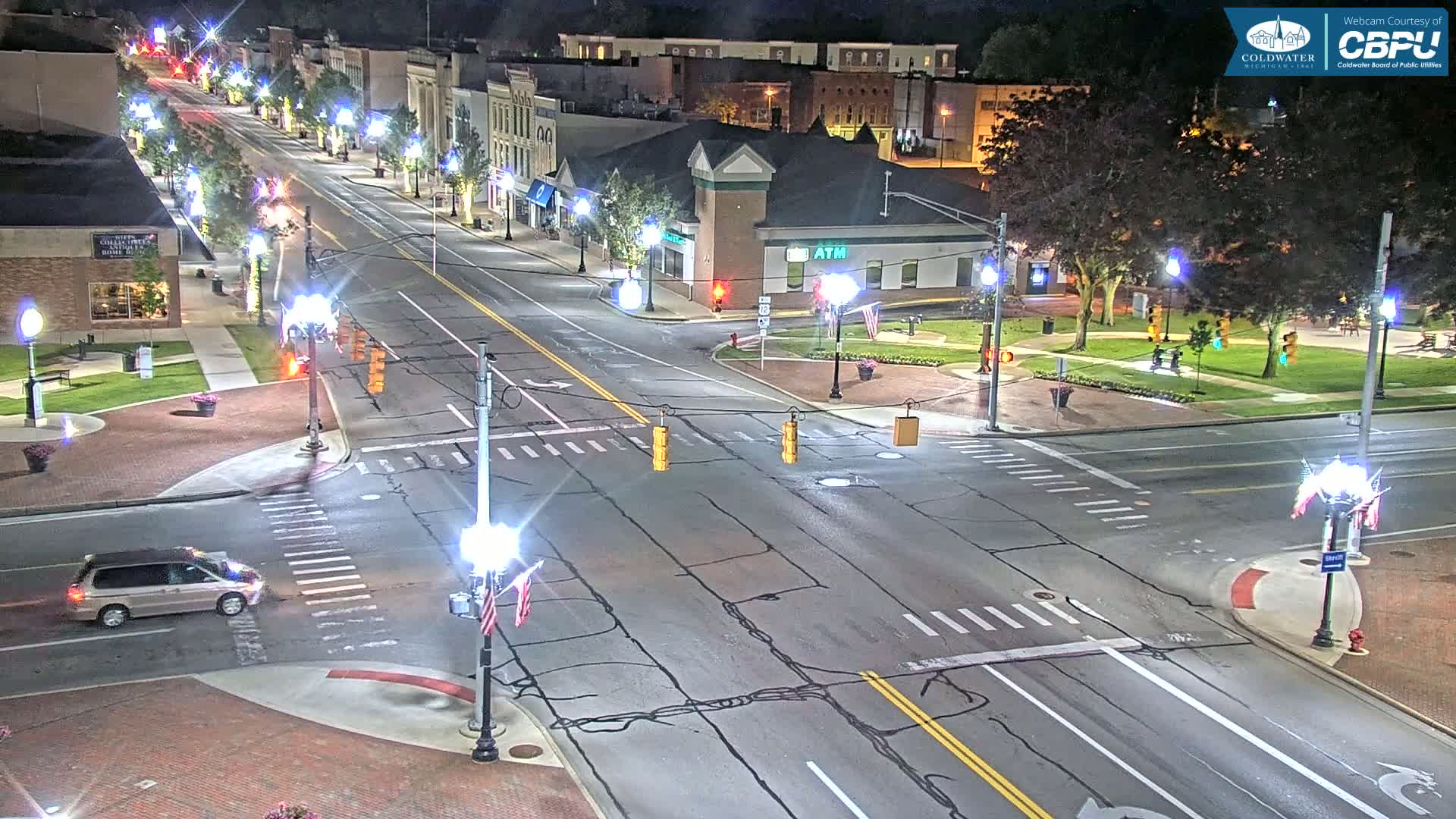 A nighttime, high-angle view of a mostly empty, four-lane road in a town, intersecting with another road at a traffic light, lined with buildings and trees.