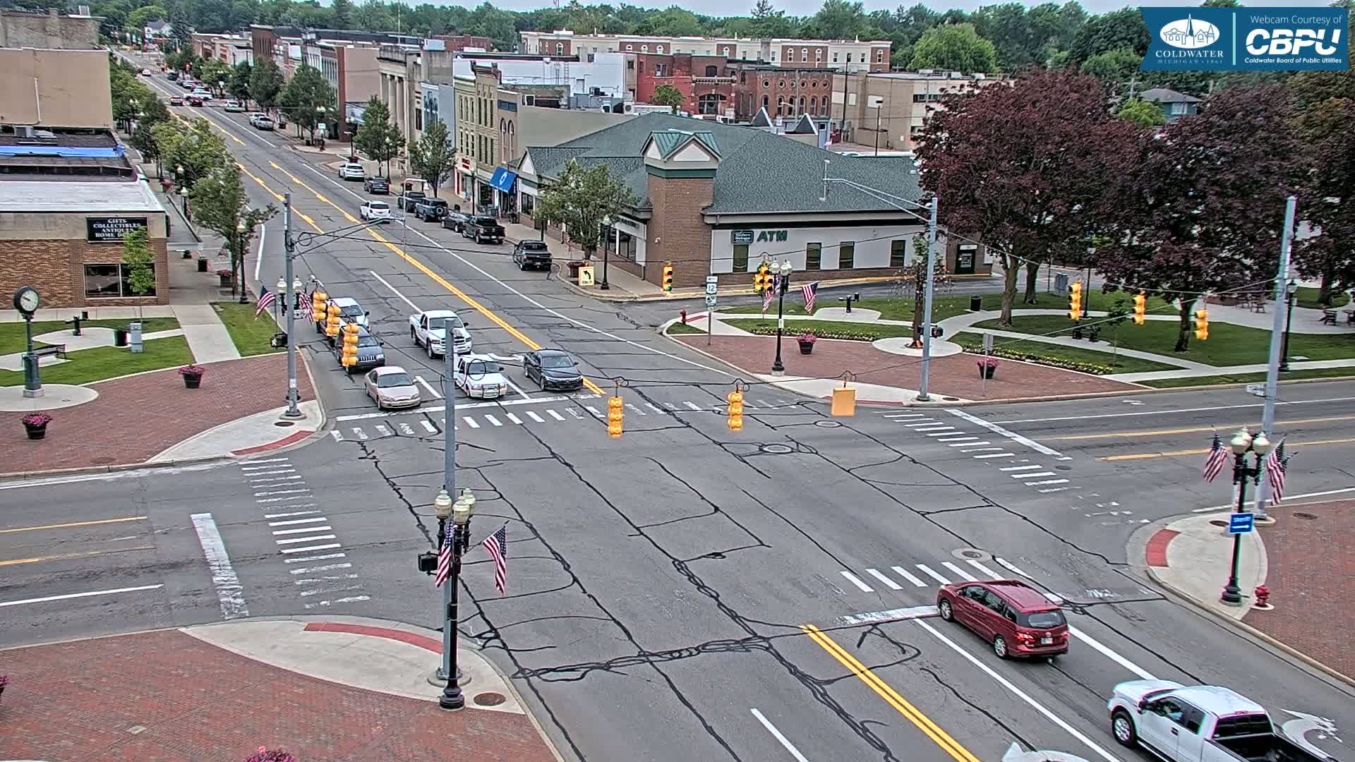 A partly sunny day shows a city intersection with several cars driving on a multi-lane road,  surrounded by brick sidewalks, landscaping, and two-story buildings.