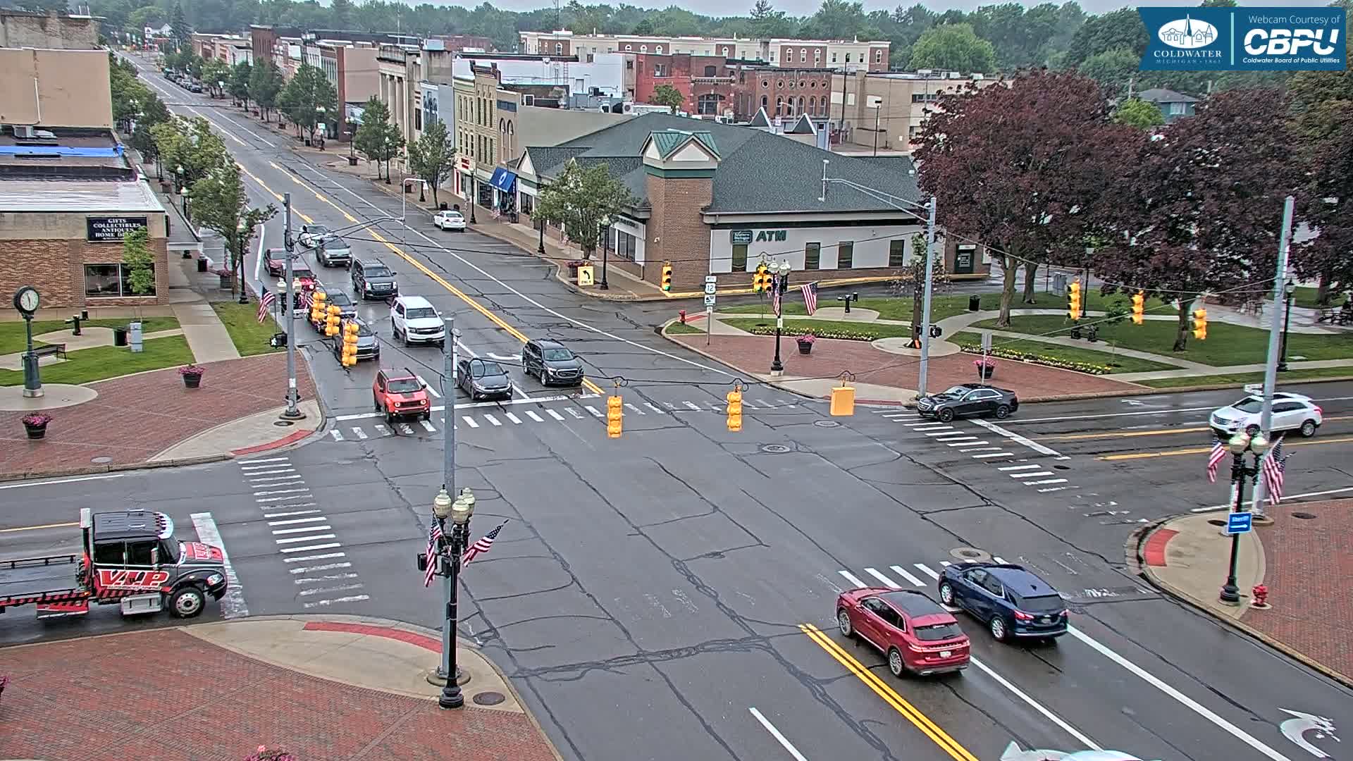 A wet city street scene shows several cars at a crossroads in front of brick buildings under an overcast sky.
