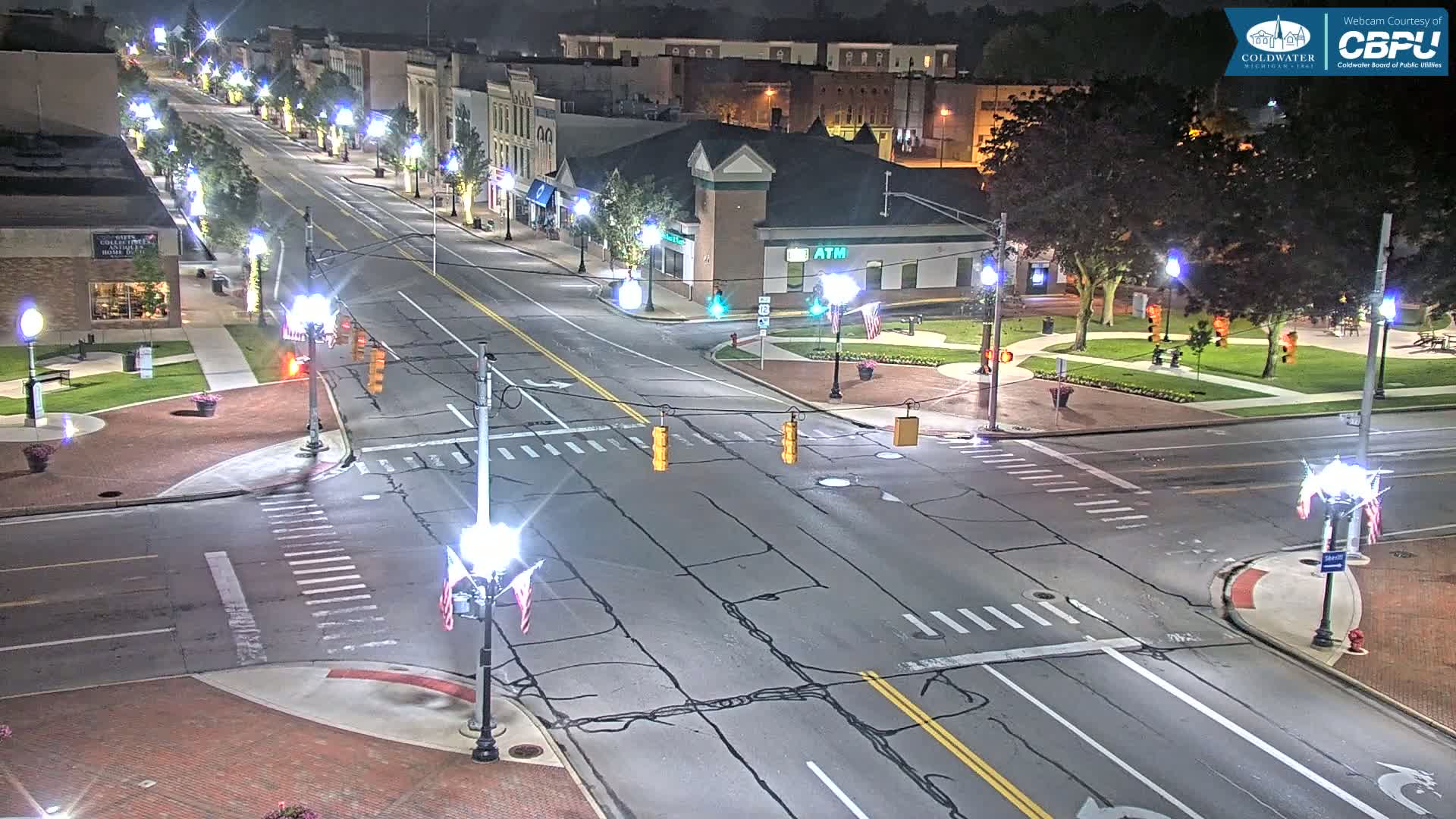 A nighttime aerial view of a town square intersection with traffic lights and streetlights illuminating a mostly empty street lined with buildings.