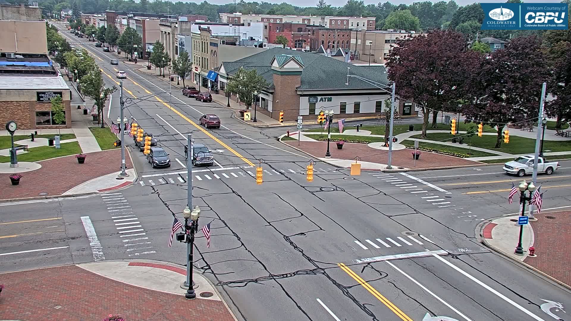 A mostly sunny day shows a street intersection in a town with several cars driving through, lined with buildings and trees, and small American flags decorating light posts.