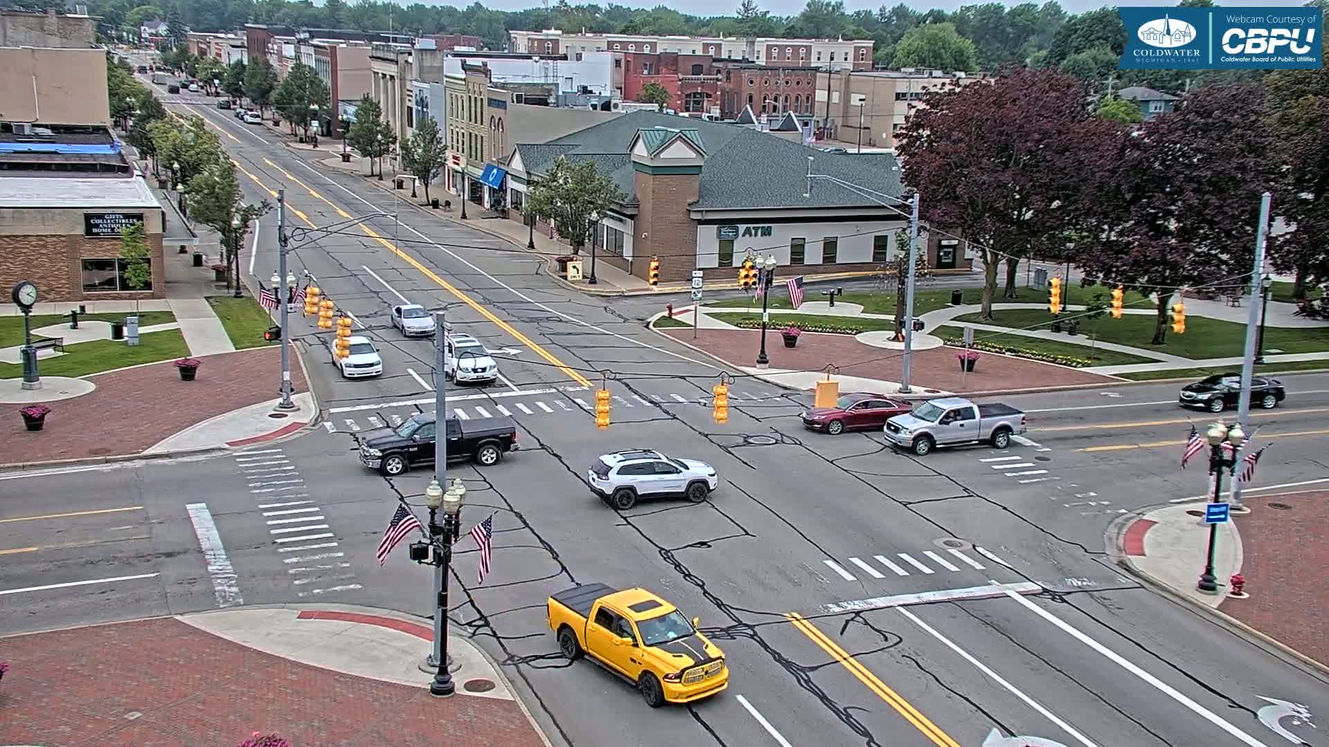 A sunny day view of a town's intersection with several vehicles and American flags displayed near traffic lights, amidst brick-paved areas and buildings.