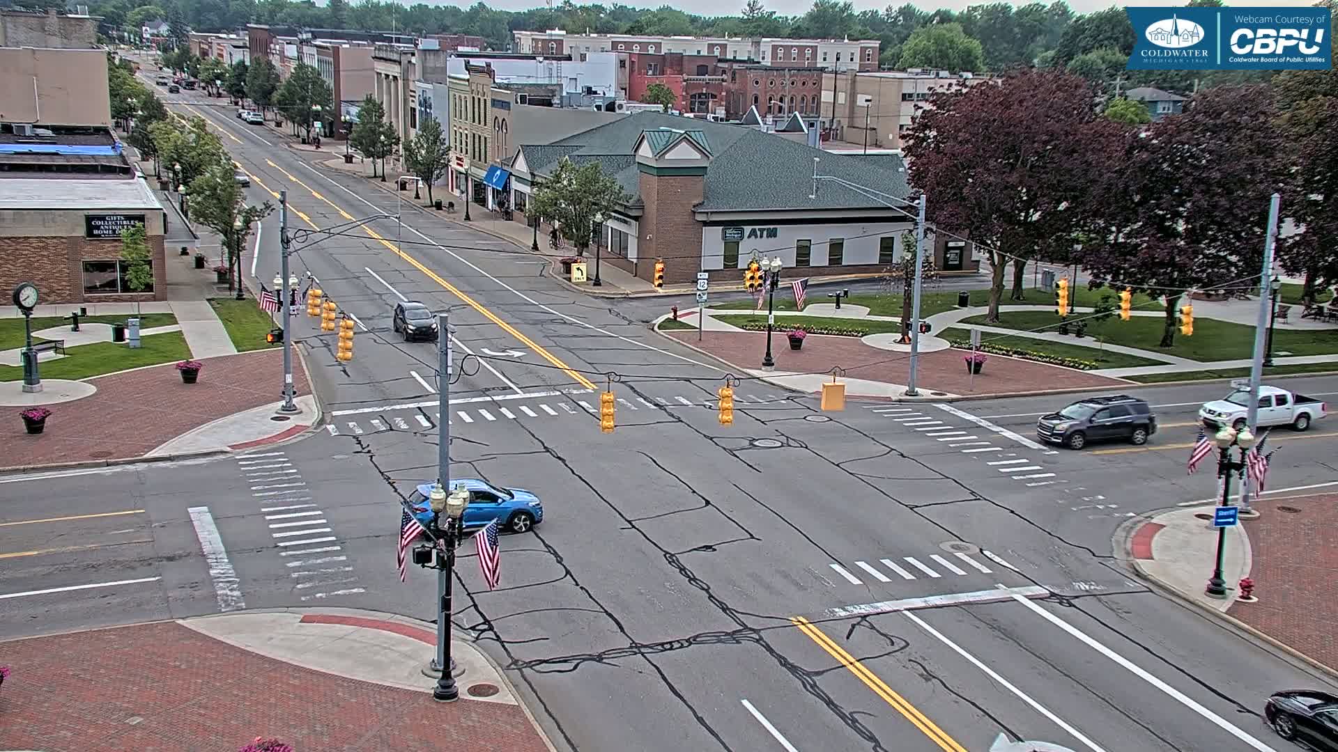 A sunny day view of a town's intersection with several vehicles and American flags displayed near traffic lights, amidst brick-paved areas and buildings.