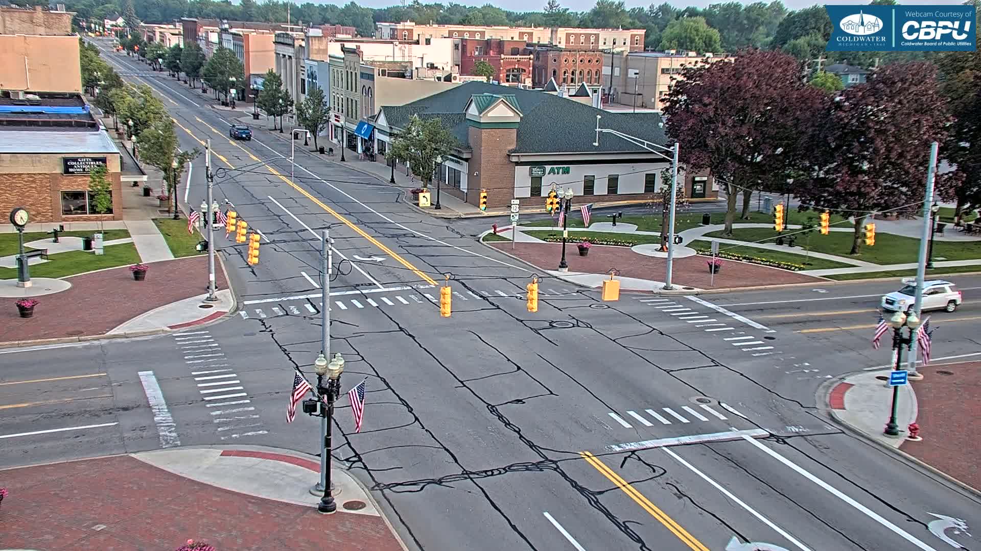 A mostly sunny overhead view of a town square intersection with several buildings, trees, and American flags, showing light traffic on the roads.