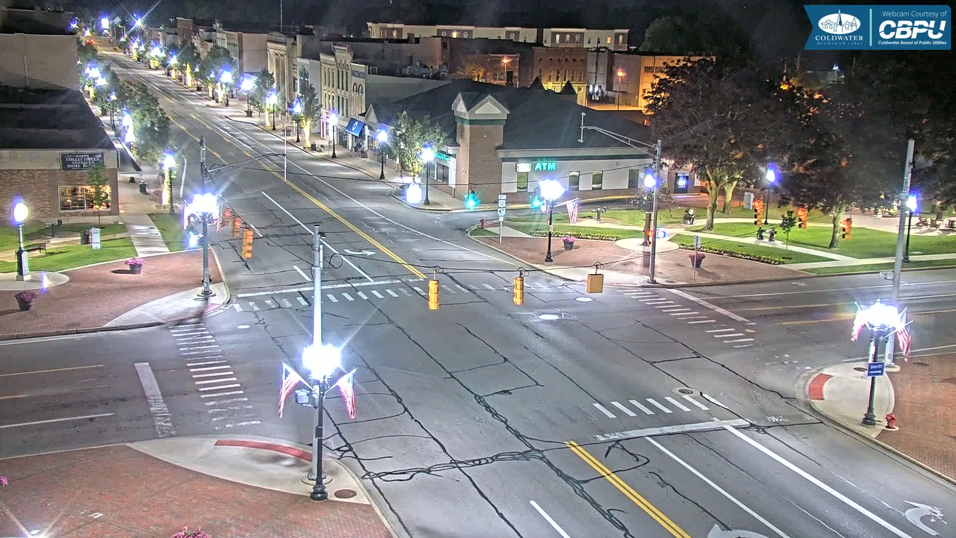 An empty, four-lane road at night intersects with another road at a crosswalk, surrounded by buildings, trees, and streetlights in a town square.