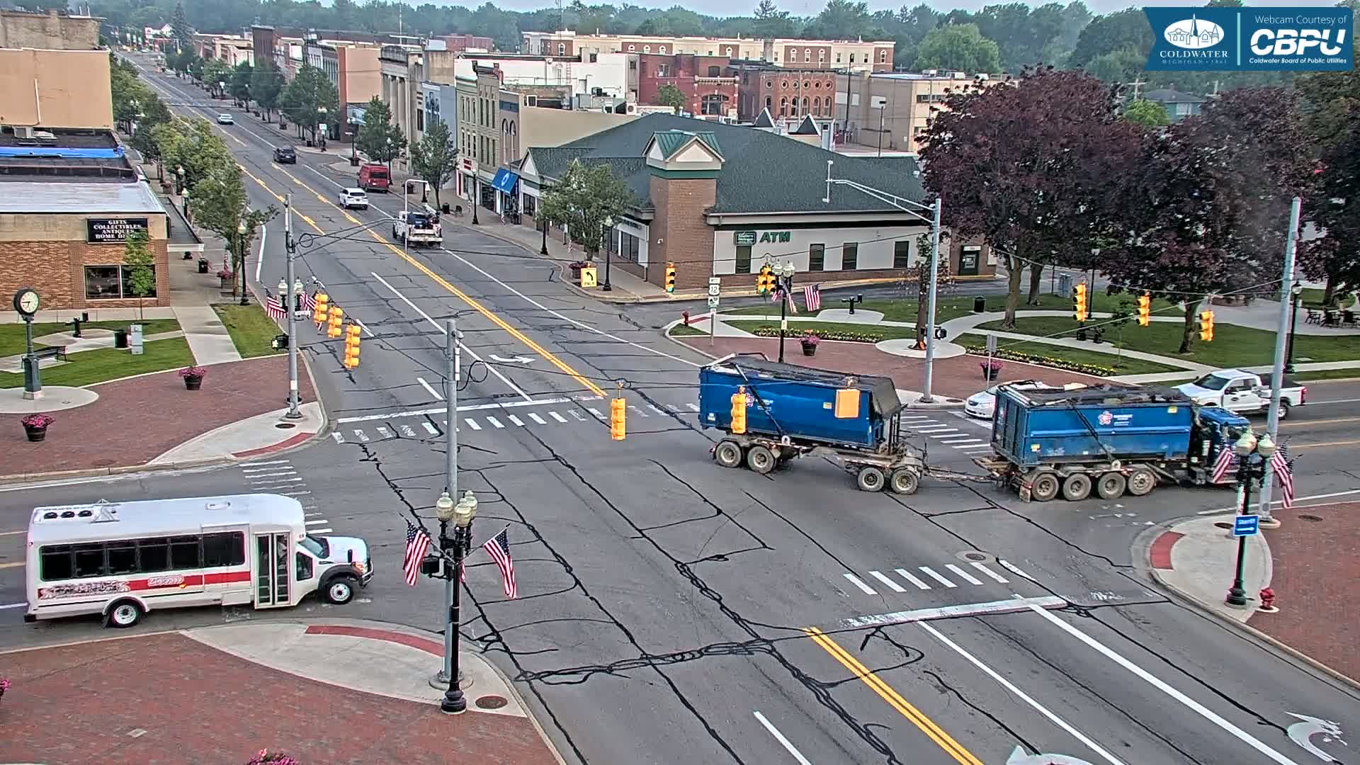 A partly sunny day shows a high-angle view of a town's intersection where two blue dump trucks and a white minibus are present along with other vehicles, and several buildings are visible in the background.