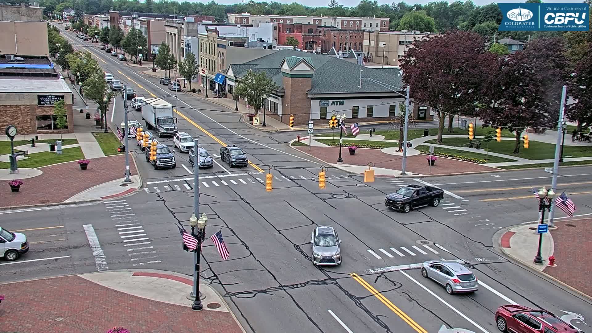 A partly sunny day shows a four-way intersection in a town with several cars driving and parked, surrounded by brick sidewalks, grass, trees, and buildings.