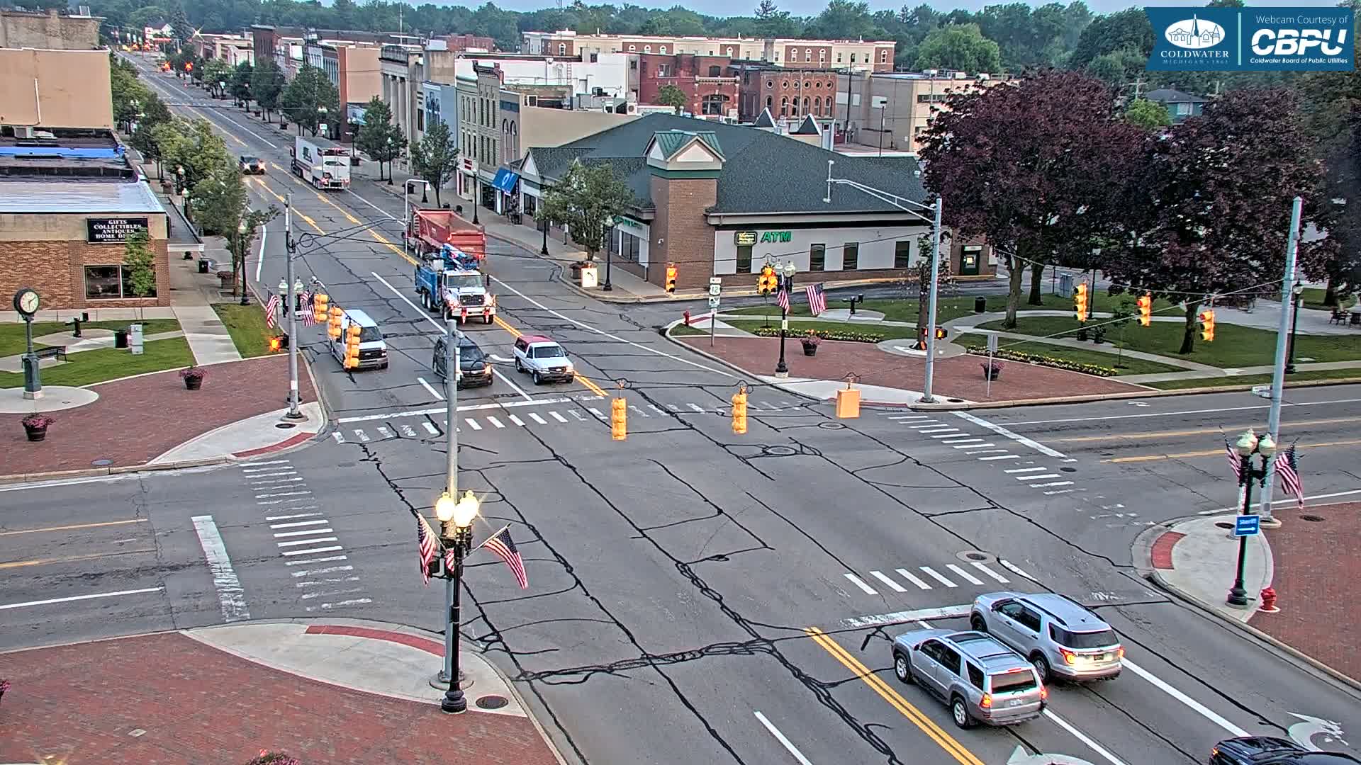 A partly cloudy daytime scene shows a multi-lane road intersection in a small town, with several vehicles driving through and several American flags displayed on light posts.