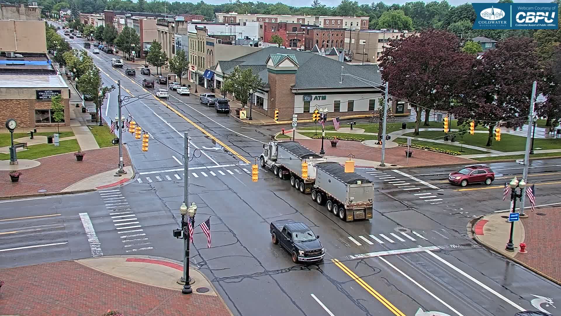A wet, tree-lined downtown street intersection shows a large truck and several smaller vehicles, with buildings in the background.