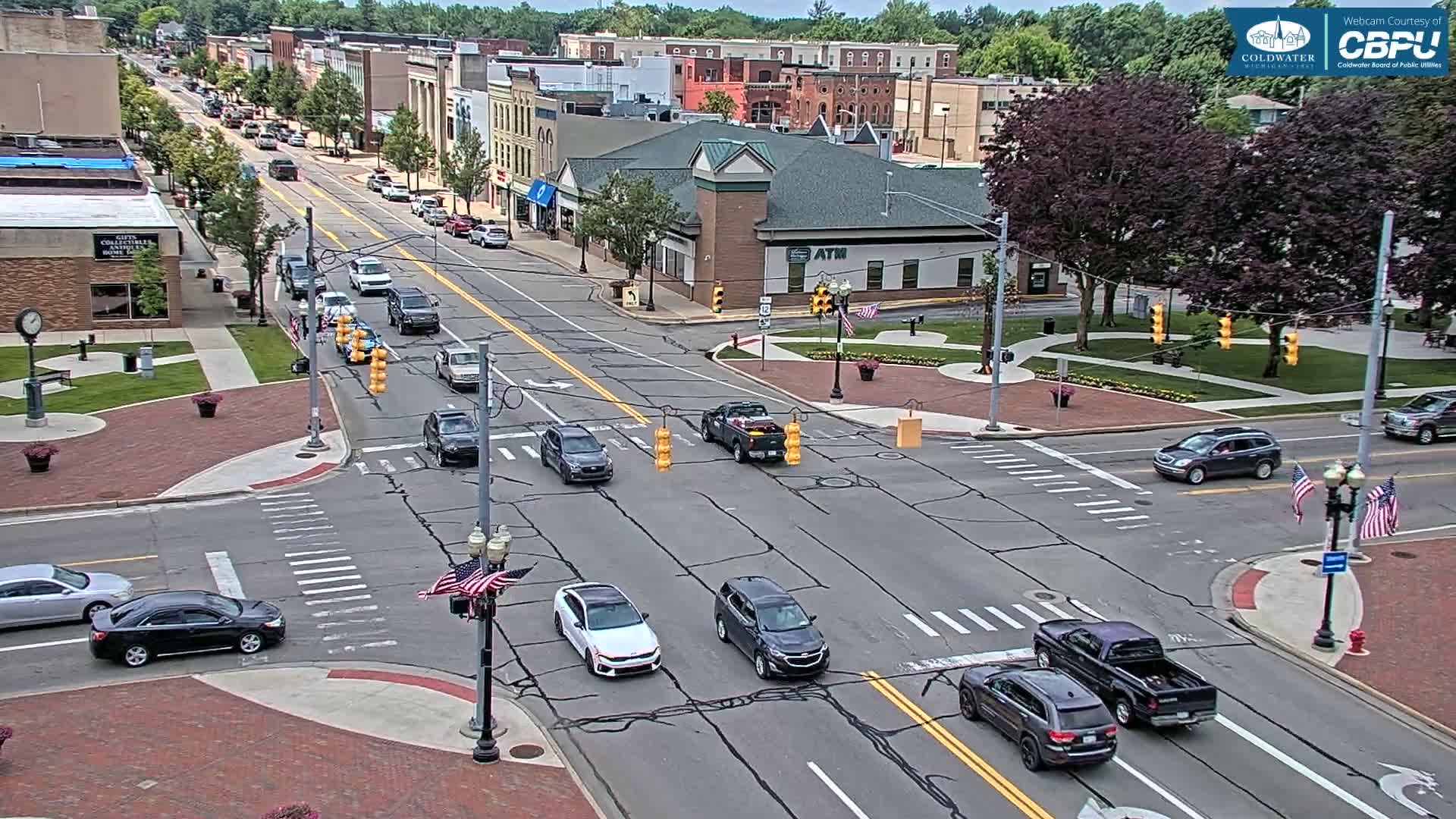 A sunny day reveals a bird's-eye view of a town's intersection with several cars moving through it, surrounded by brick sidewalks, buildings, and lush green trees.