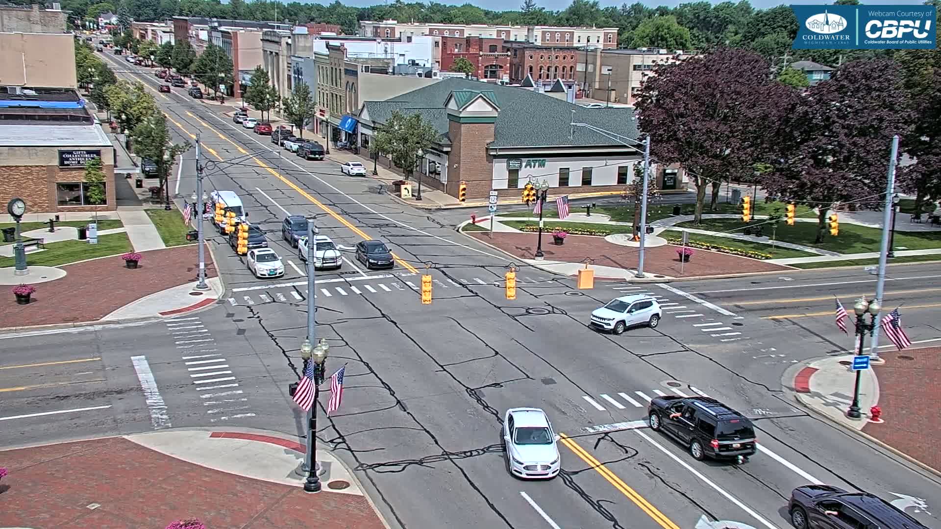 A sunny day in a town square shows several cars at a multi-way intersection with brick sidewalks, green spaces, and American flags on light poles.