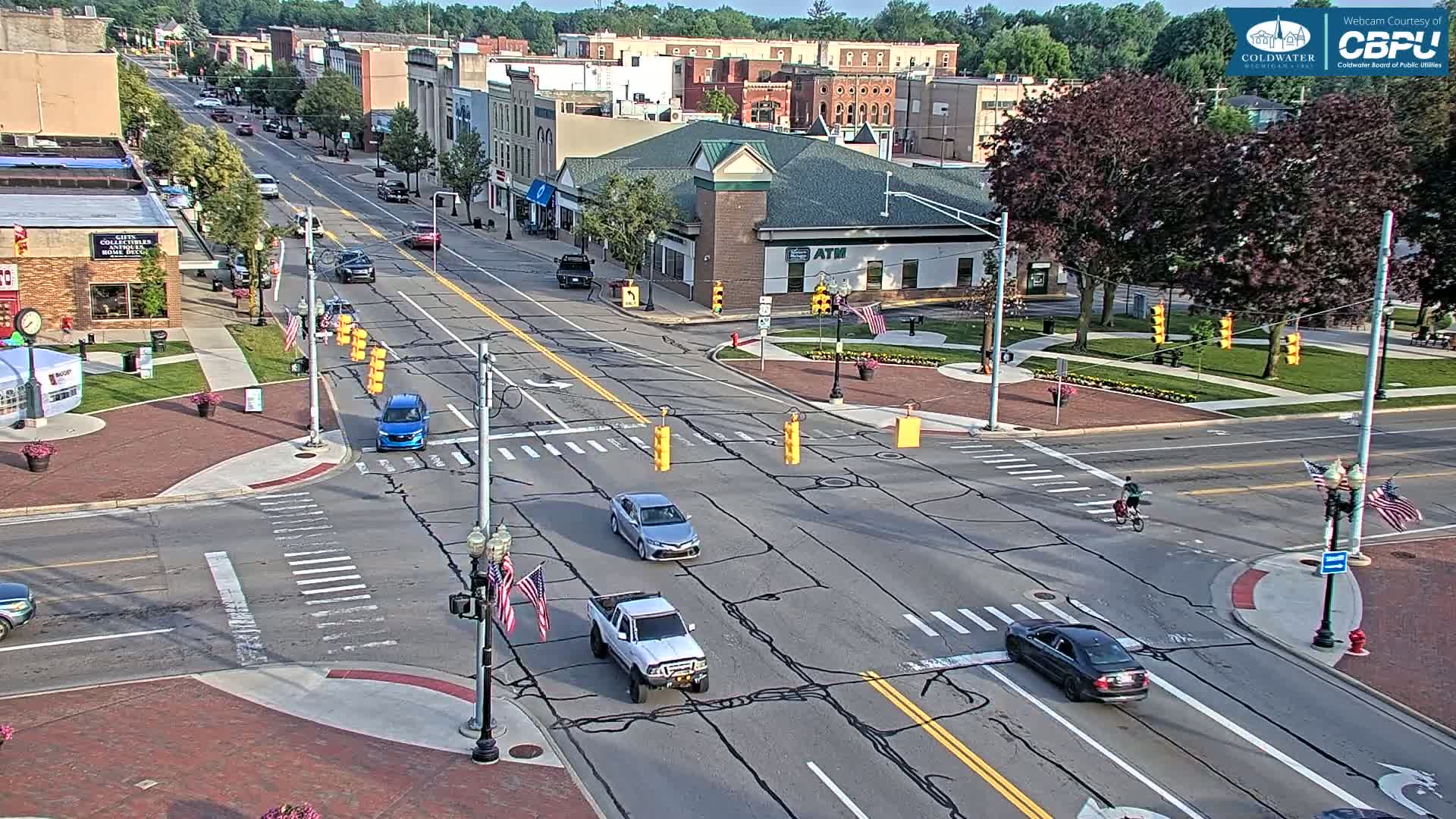 A sunny day reveals a high-angle view of a town's intersection, where several vehicles are navigating the crossroads amidst buildings and green spaces.