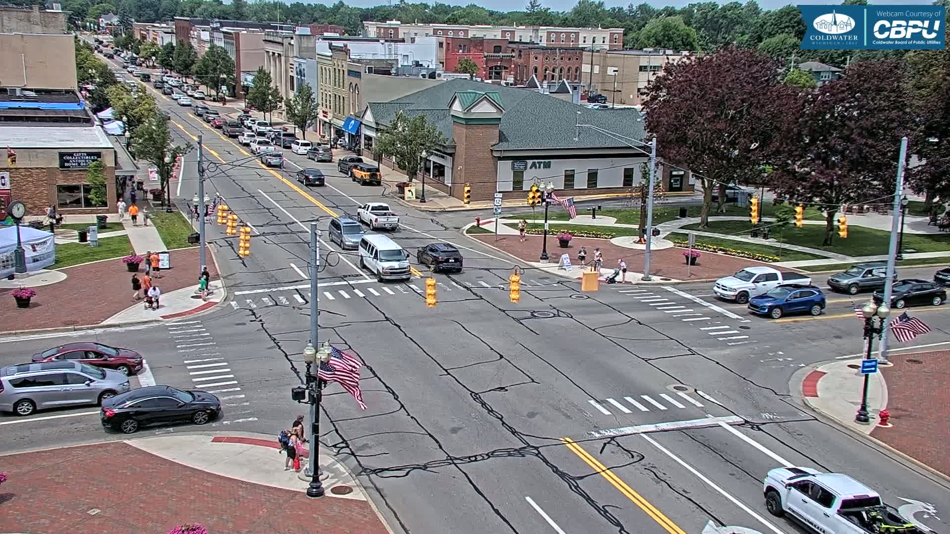 A sunny day shows a street intersection in a town with multiple cars and pedestrians, surrounded by buildings and green spaces.