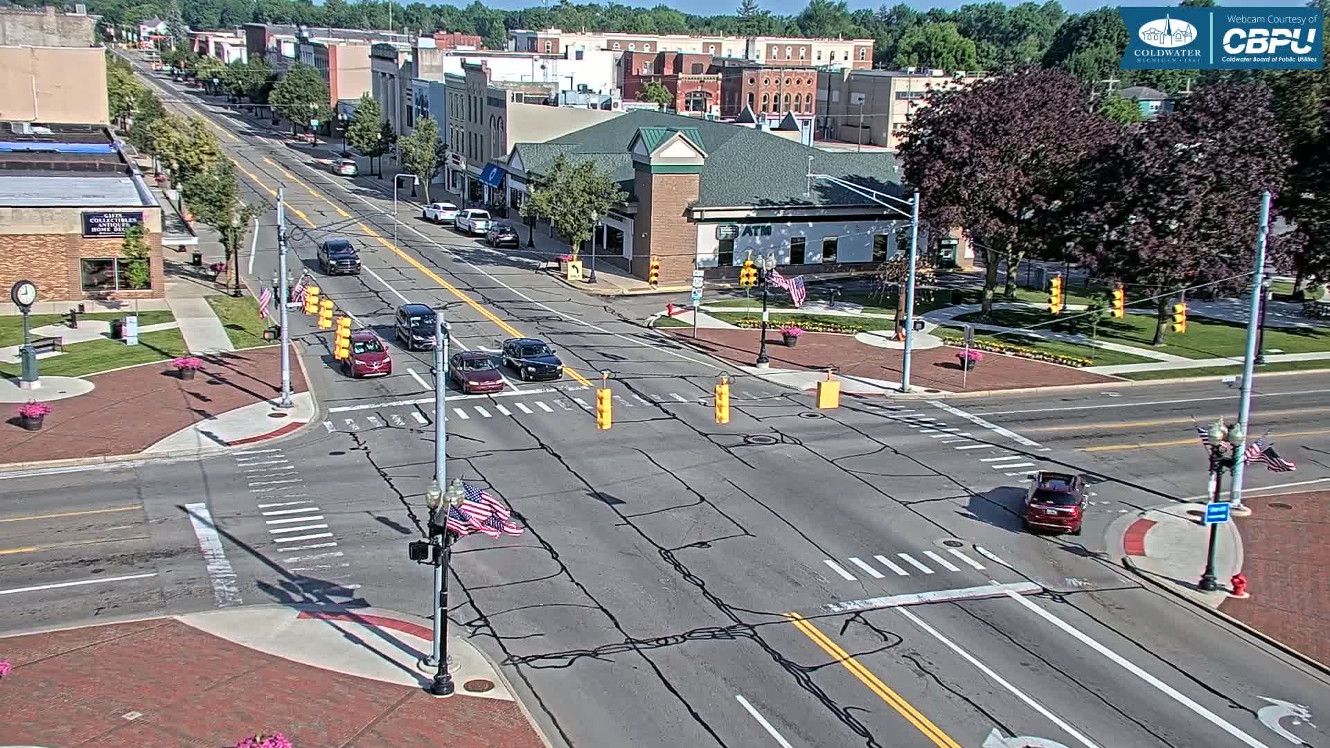 A sunny day reveals a town's main street intersection with several cars and a few pedestrians, surrounded by brick buildings and green spaces.