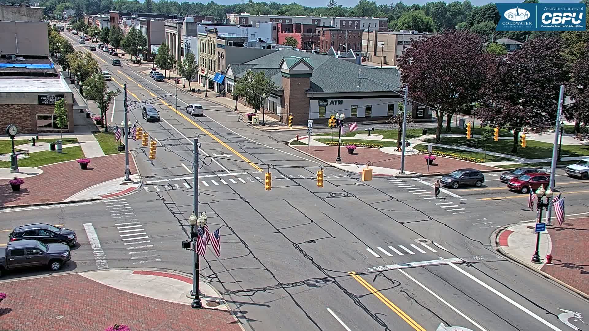 A high-angle view of a city intersection on a sunny day, showing several cars at a crosswalk and pedestrians crossing the street.
