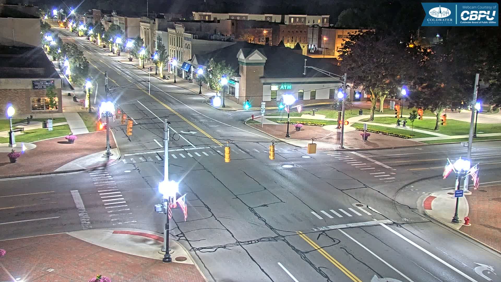 A mostly empty, well-lit street intersection in a town at night, with buildings lining the street and trees in a nearby park.